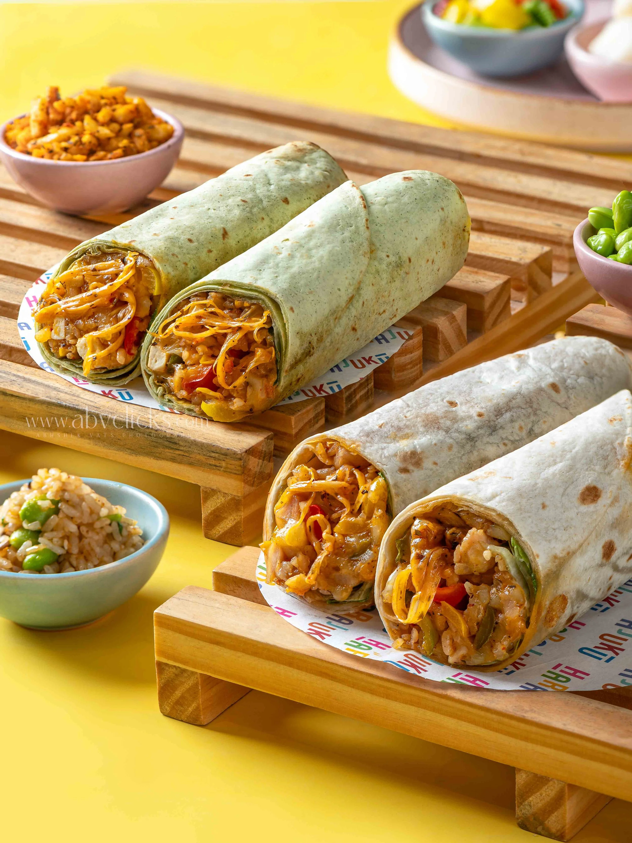 Three burritos filled with rice, vegetables, and cheese sit on a wooden tray, with small bowls of rice, salad, and green beans around them, set against a bright yellow background.