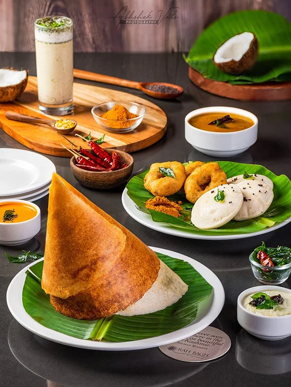 An Indian meal featuring a large crispy dosa on a green banana leaf, onion bhajis, and idlis garnished with herbs, along with bowls of sambar, chutney, and a coconut.