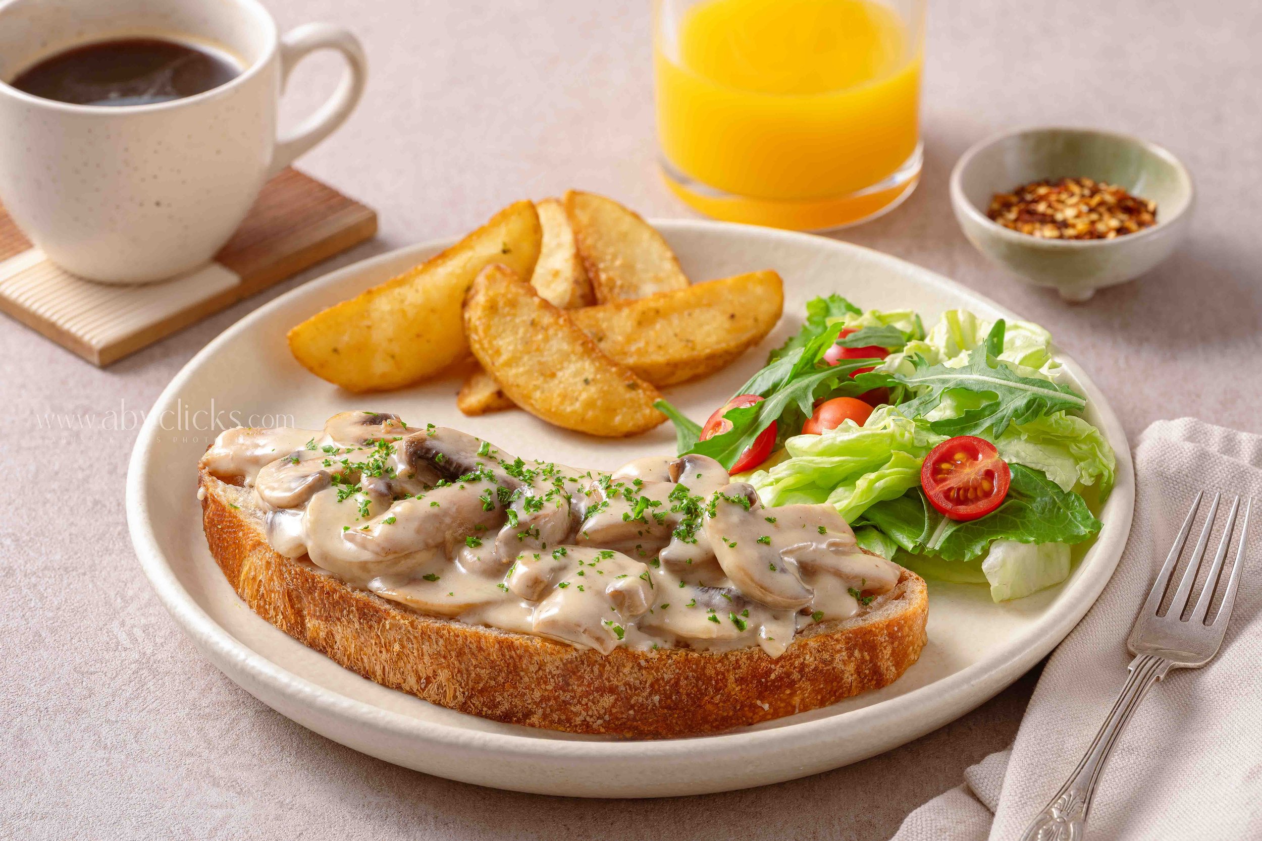 A breakfast plate featuring toast with mushroom gravy, a side salad with cherry tomatoes and arugula, potato wedges, a glass of orange juice, a cup of coffee, and a small bowl of red pepper flakes.