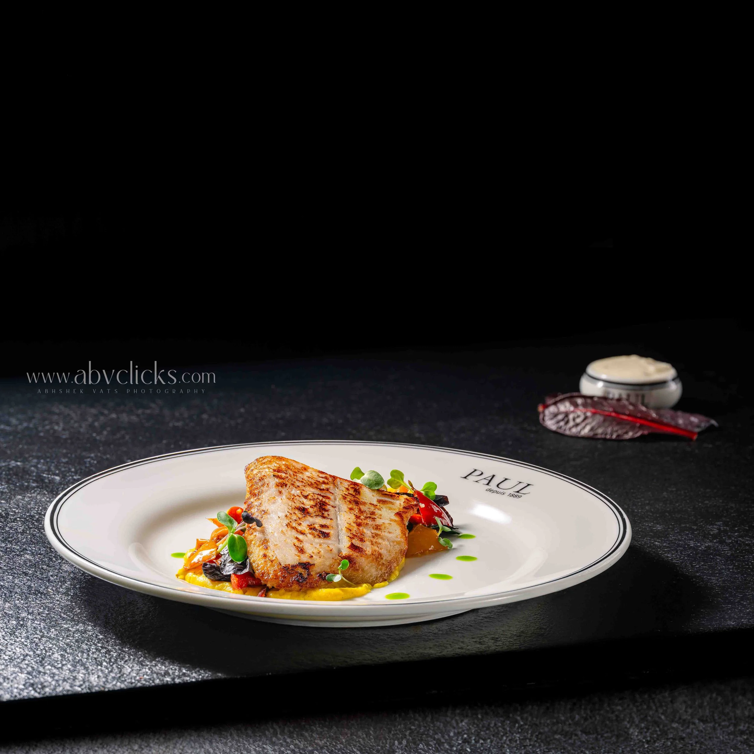 A plated gourmet dish consisting of a seared piece of fish served with vegetables on a white plate with a decorative black border, set on a dark surface.