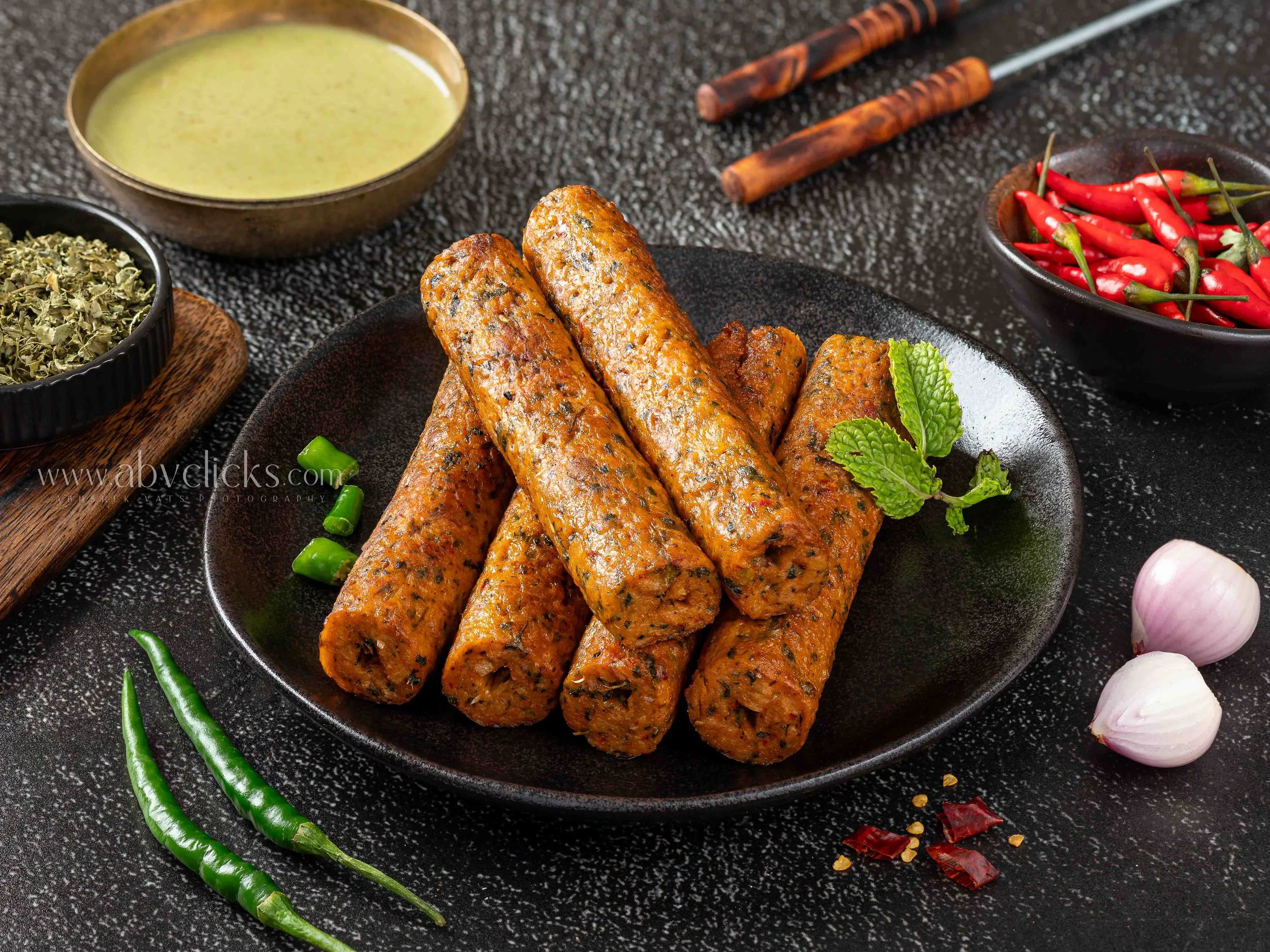 Close-up of spiced sausage rolls on a black plate, garnished with fresh mint leaves, with chili peppers, garlic, and spices on a black surface background.