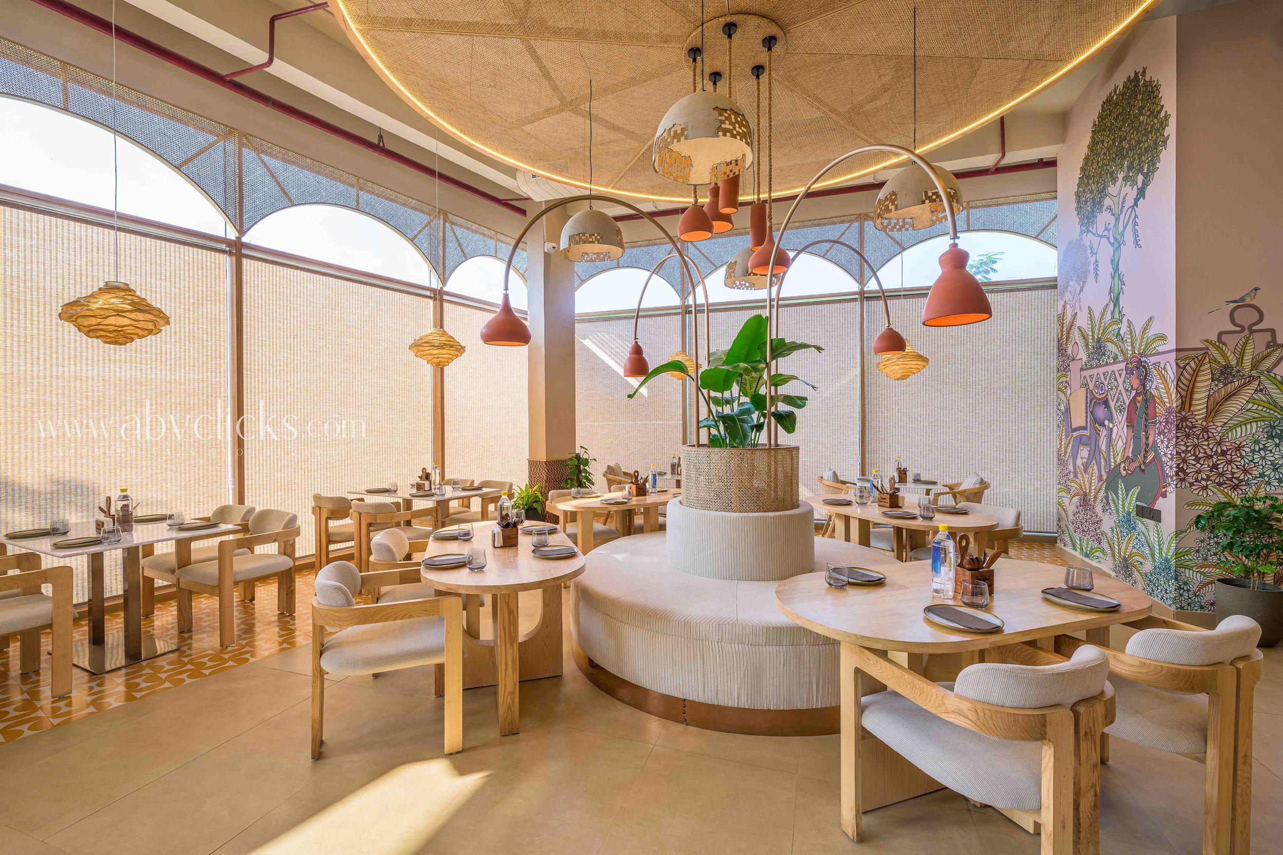 Interior of a modern restaurant with round wooden tables, beige cushioned chairs, and decorative lighting fixtures hanging from the ceiling. The restaurant features a colorful mural on the wall and large windows with shades allowing natural light to 