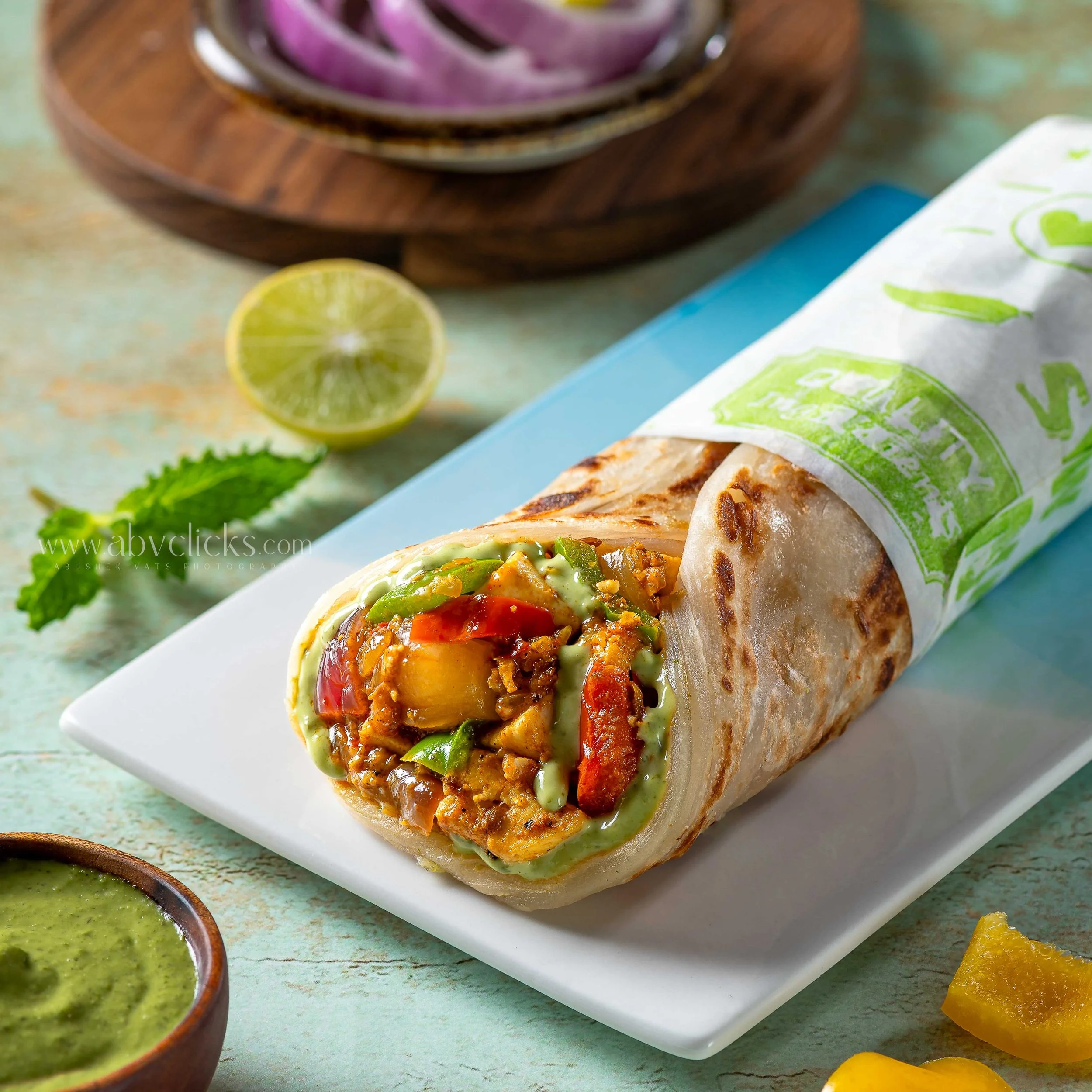 Close-up of a paneer tikka wrap with colorful bell peppers and sauce, served on a white plate with two small bowls of green chutney and a sliced lime nearby, on a light green textured surface.