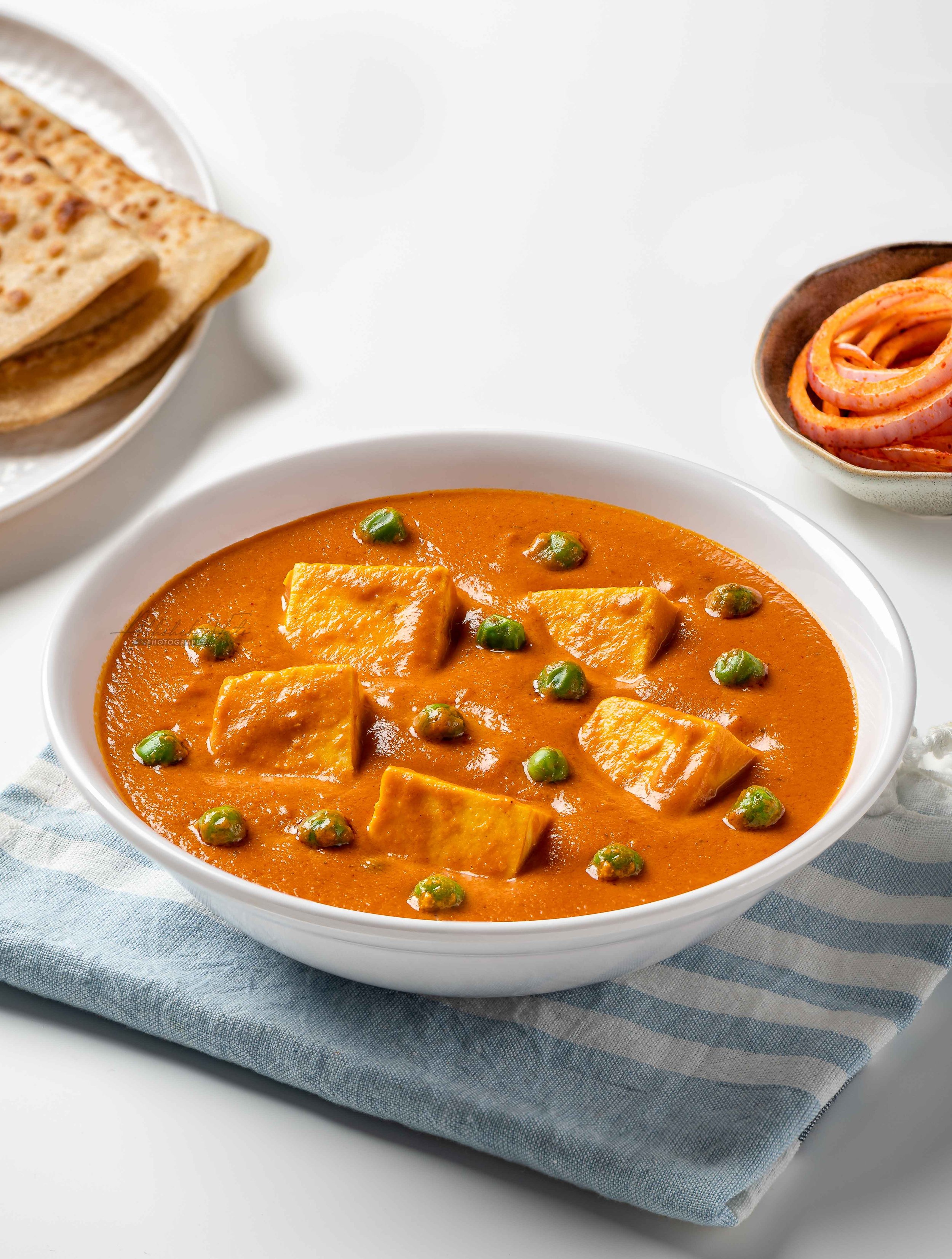 A bowl of Indian curry with chunks of paneer cheese and green peas, placed on a gray and white checkered cloth.