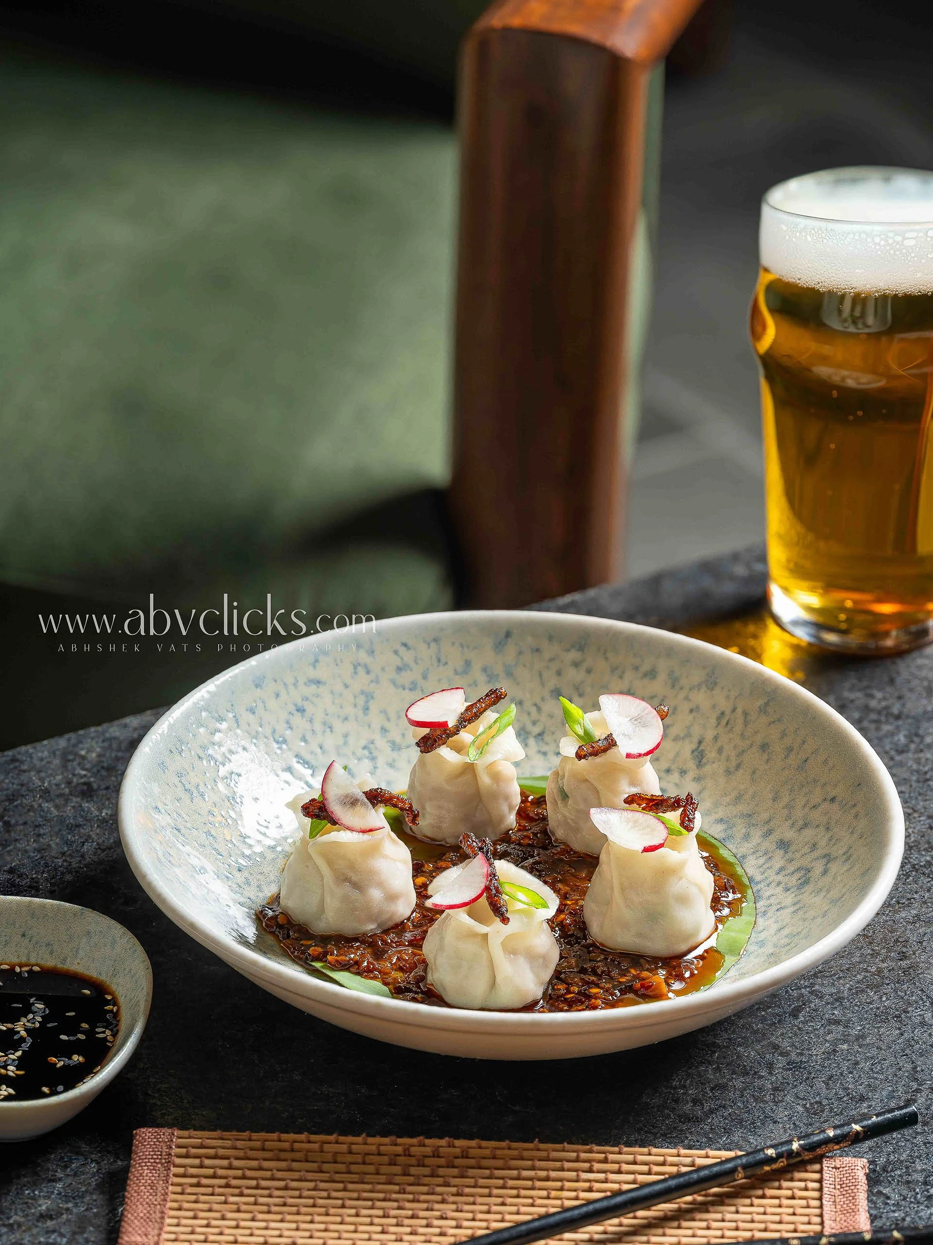 A bowl of dumplings garnished with thinly sliced radish, green onions, and fried shallots, served with a soy-based dipping sauce and a glass of beer in the background.