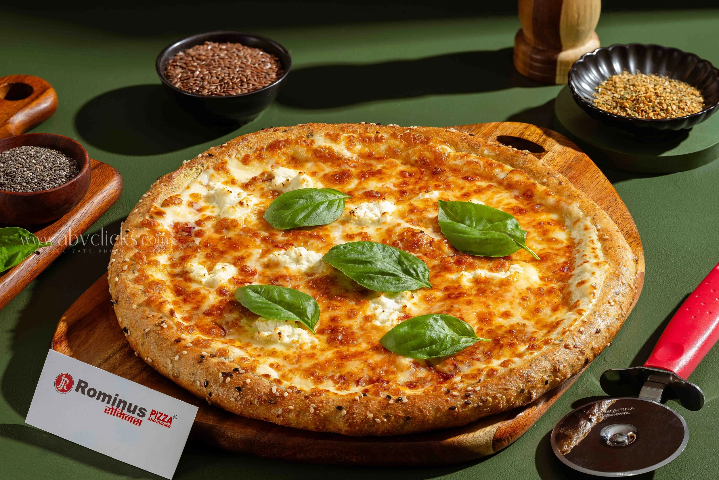 Close-up of a cheese pizza on a wooden board garnished with fresh basil leaves, surrounded by bowls of different grains and spices on a green surface.