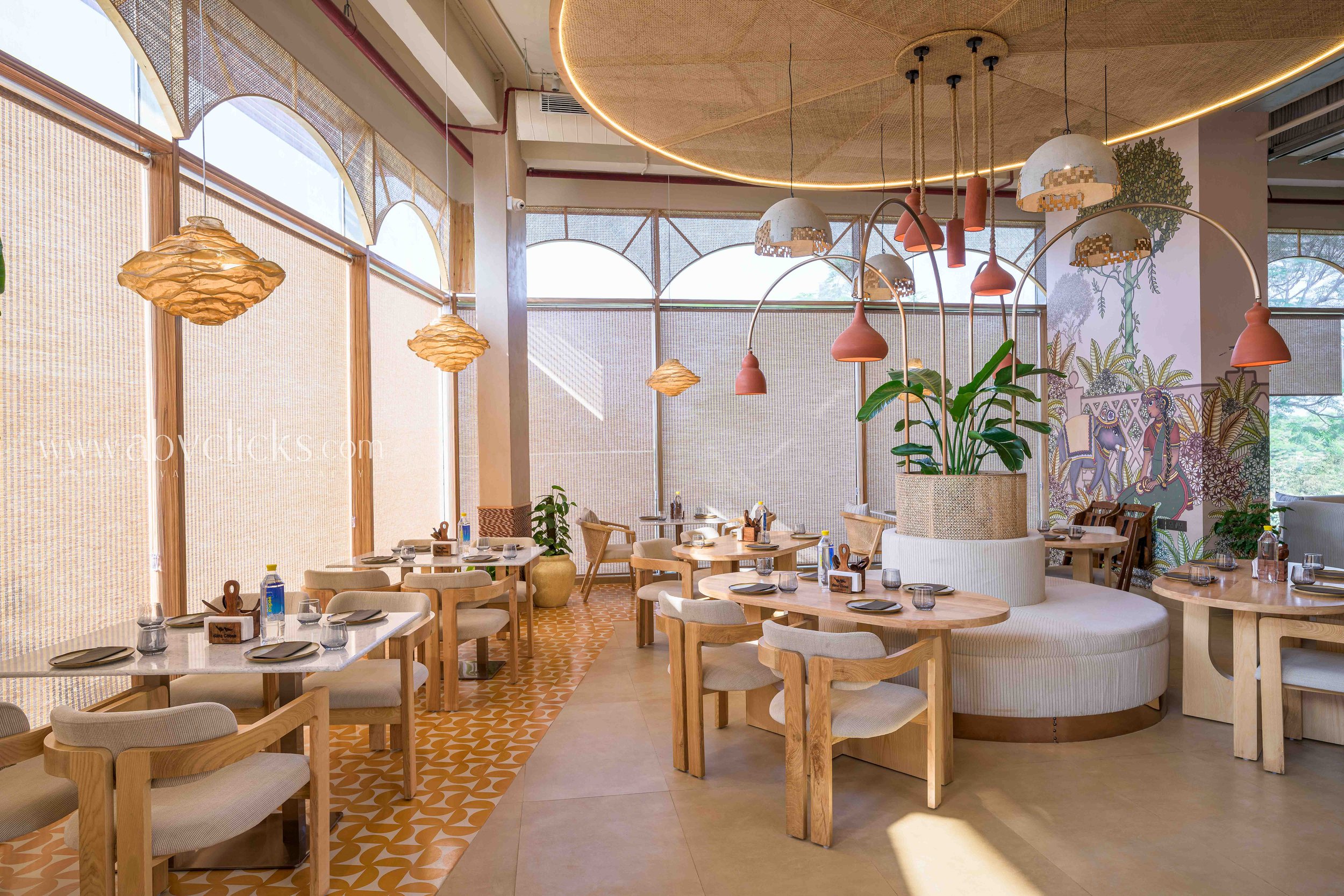 Interior of a modern restaurant with wooden tables and chairs, decorated with plants and artistic hanging light fixtures. The space features large windows, natural light, and artistic wall murals.