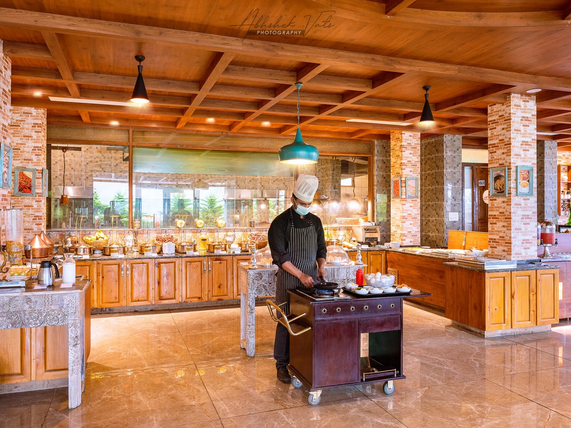 A chef wearing a face mask and a chef's hat, standing in a modern restaurant kitchen, cooking on a small cart with various bowls and ingredients, with a wooden ceiling and brick pillars in the background.