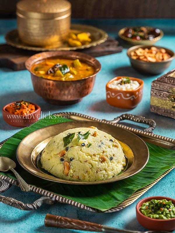 Indian meal with mashed potatoes garnished with curry leaves, served on a traditional metal plate with various bowls of curry, chutney, and spices in the background.