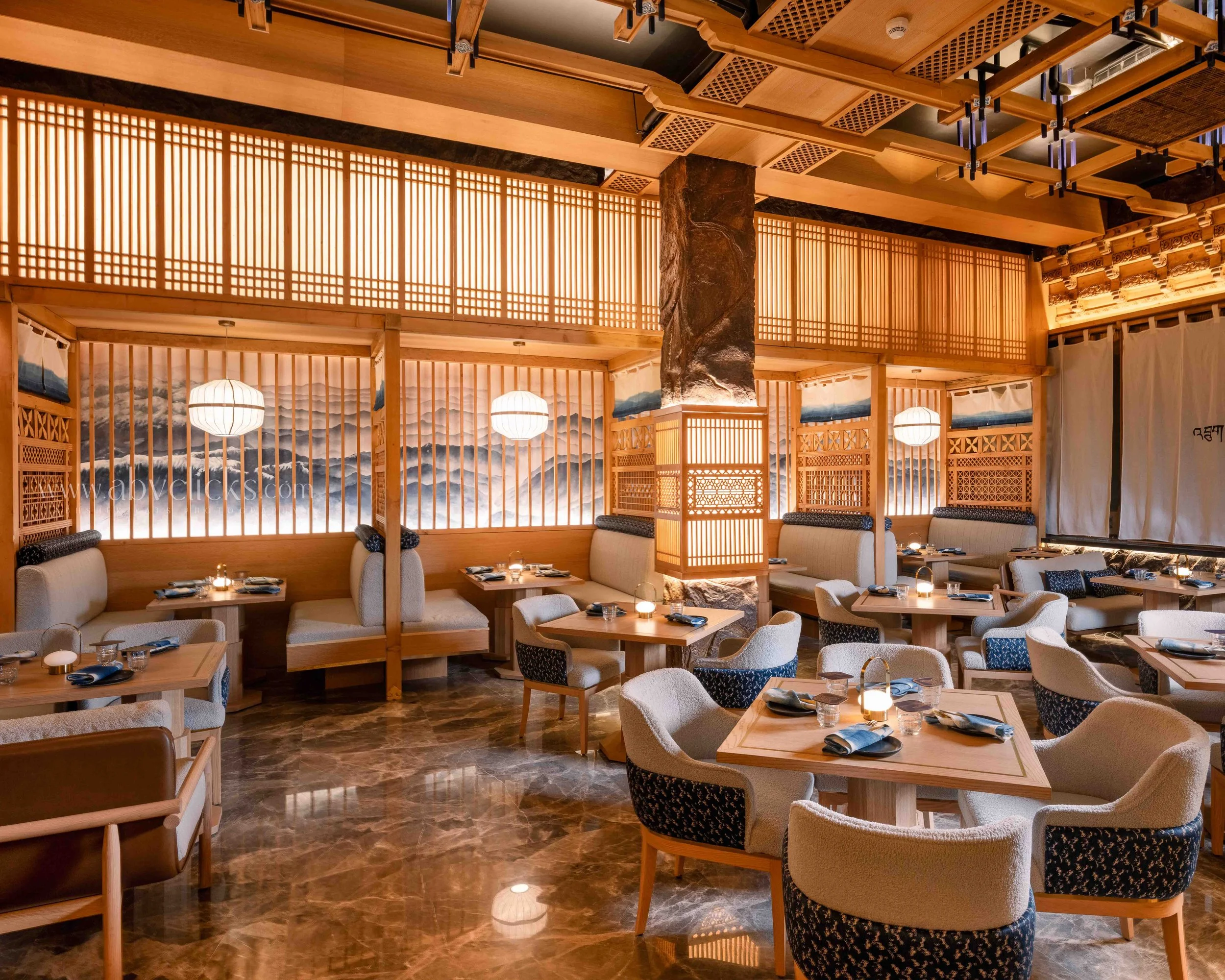 Interior of a restaurant with wooden decor, hanging lanterns, and cushioned chairs. The scene includes a painted mountain landscape on the wall and marble flooring.