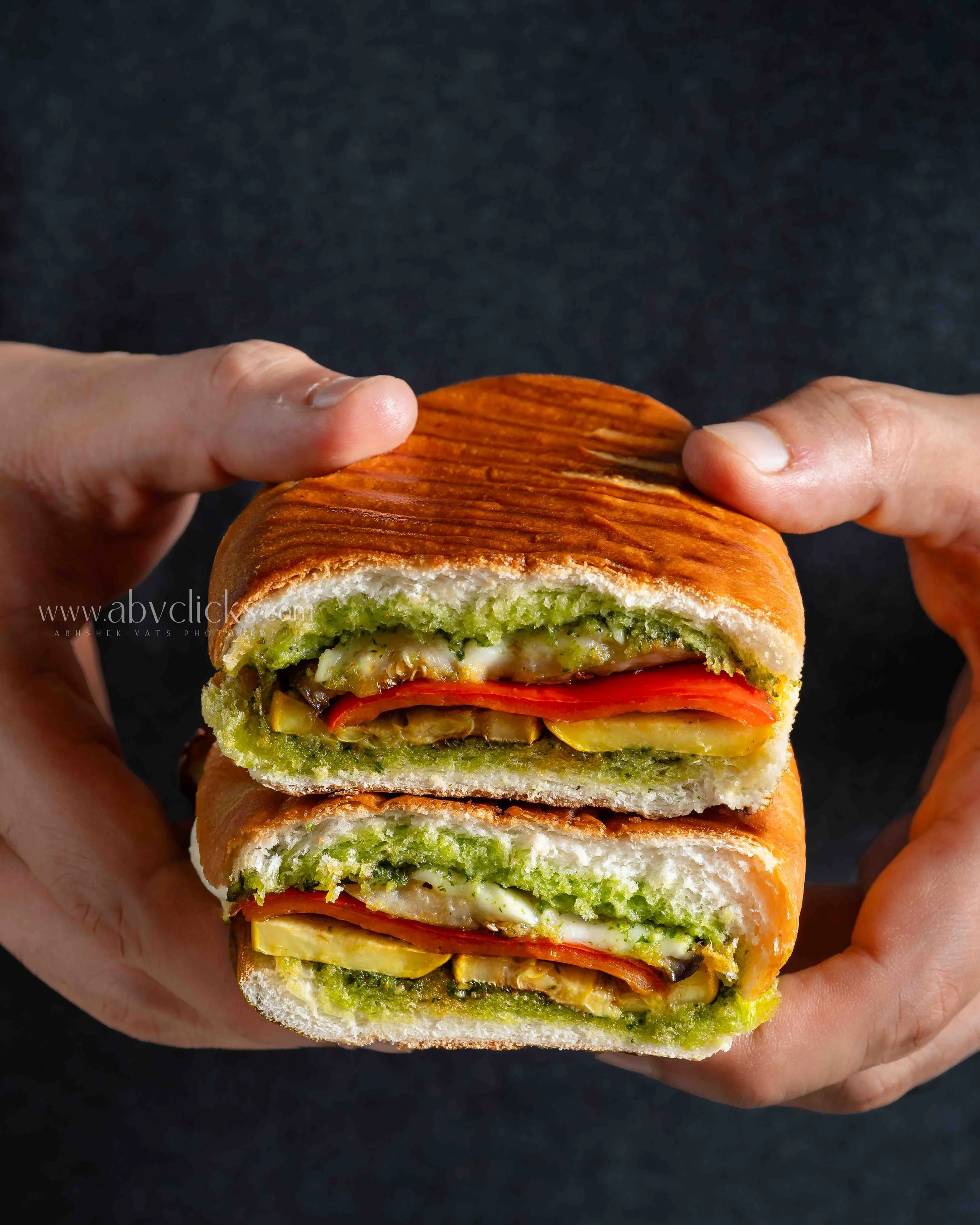 Close-up of a person's hand holding a multi-layered sandwich with vegetables, cheese, and meat, against a black background.