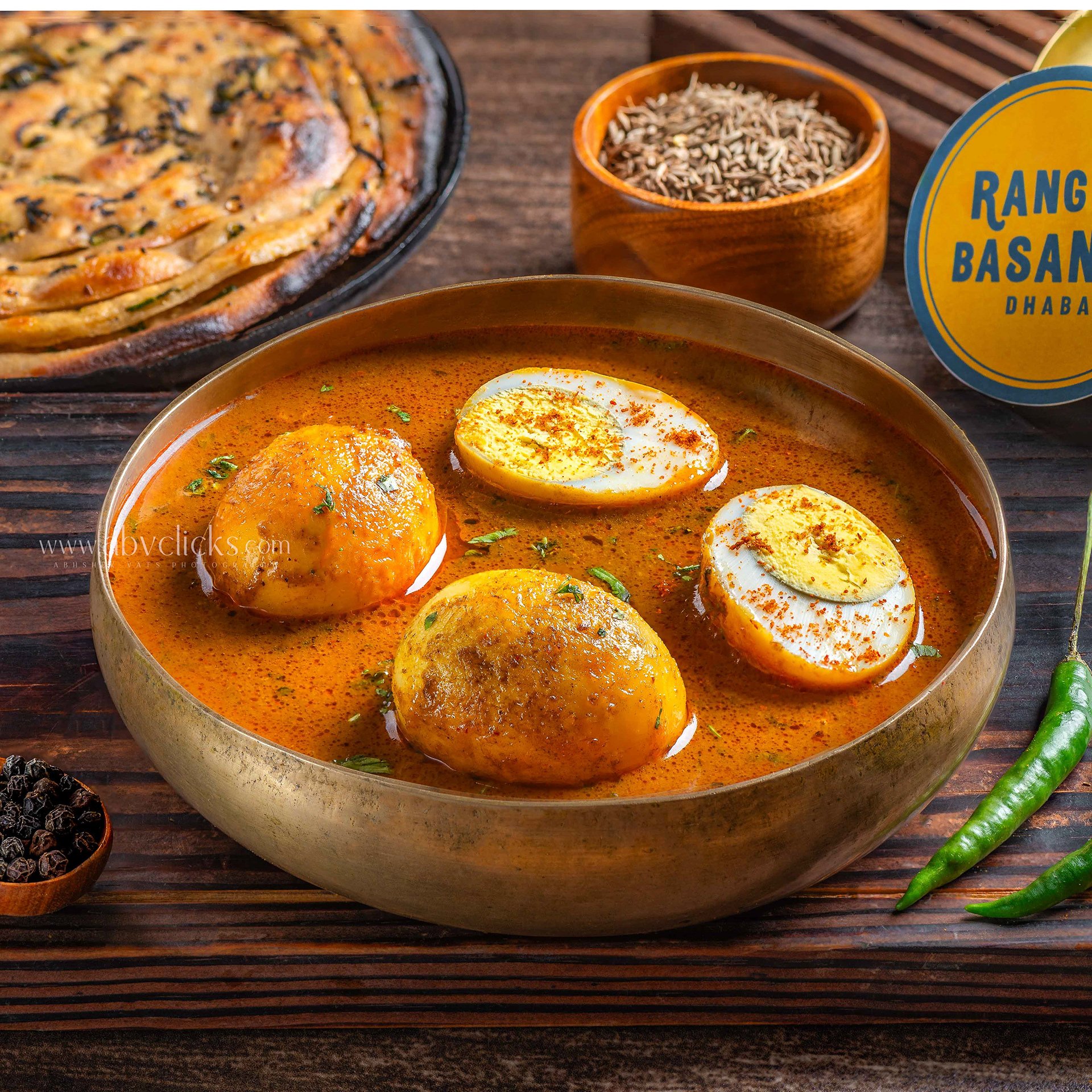 Close-up of Indian dishes including a bowl of spicy curry with boiled eggs, a plate of layered flatbread with herbs, a small bowl of black peppercorns, and a container of cumin seeds on a wooden table.