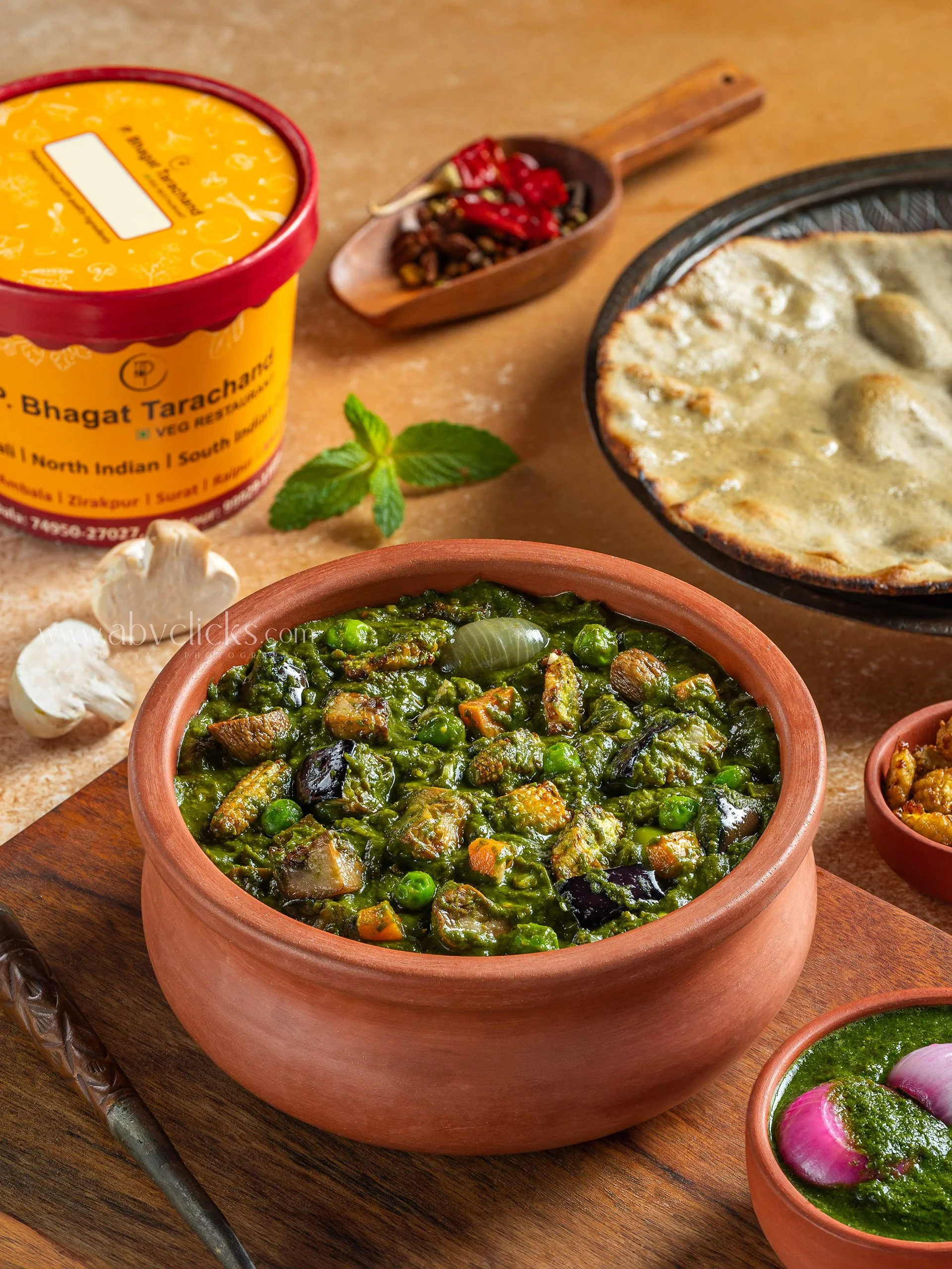 A traditional Indian vegetarian meal with a clay pot of mixed vegetable curry, a bowl of palak (spinach) sabzi, a bowl of green chutney, and a bowl of pickled onions and herbs. There is also a pizza on a plate in the background, along with dried red 