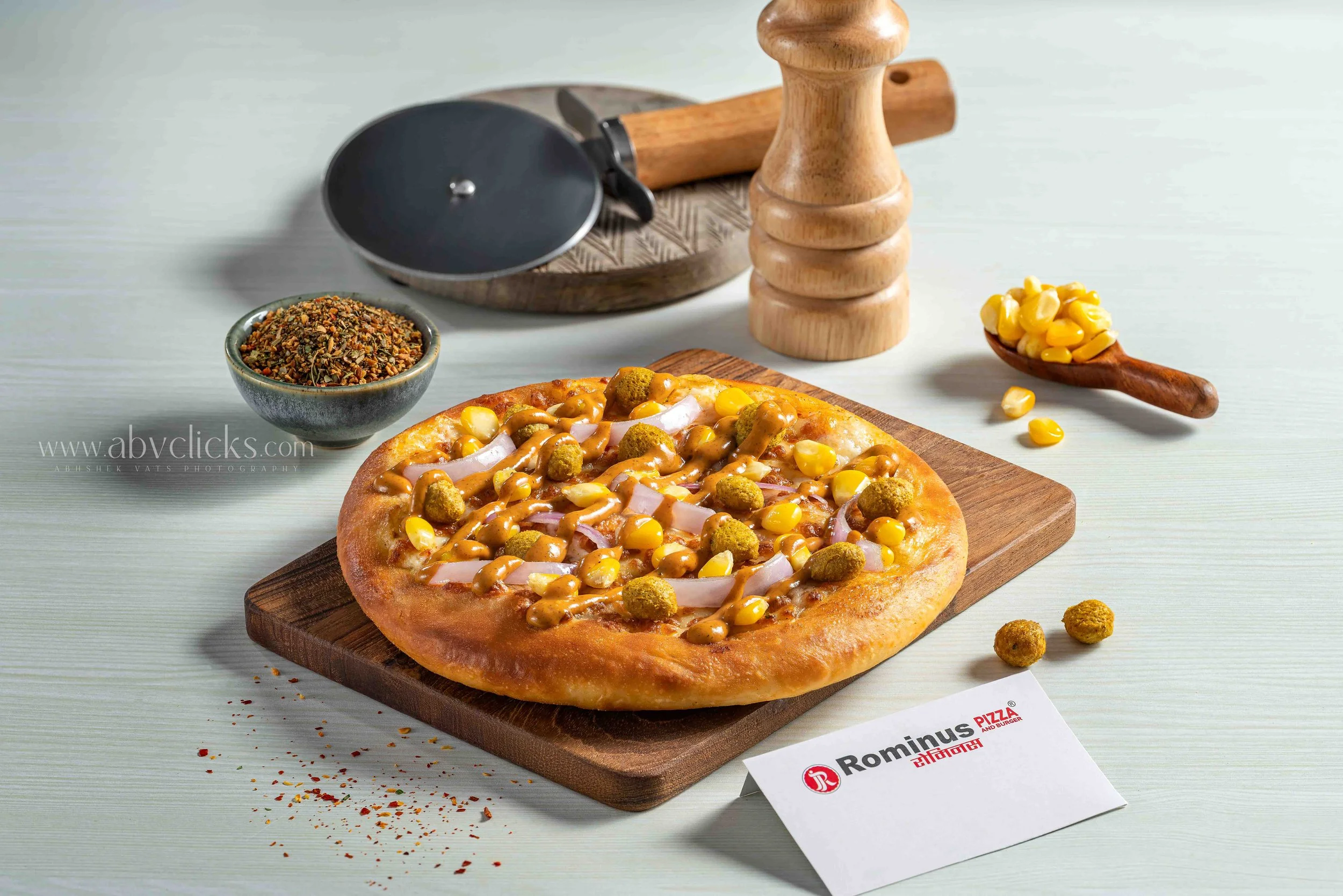 Close-up of a pizza with corn, chicken, and sauce on a wooden board, with condiments, spices, and a restaurant name card nearby on a white surface.