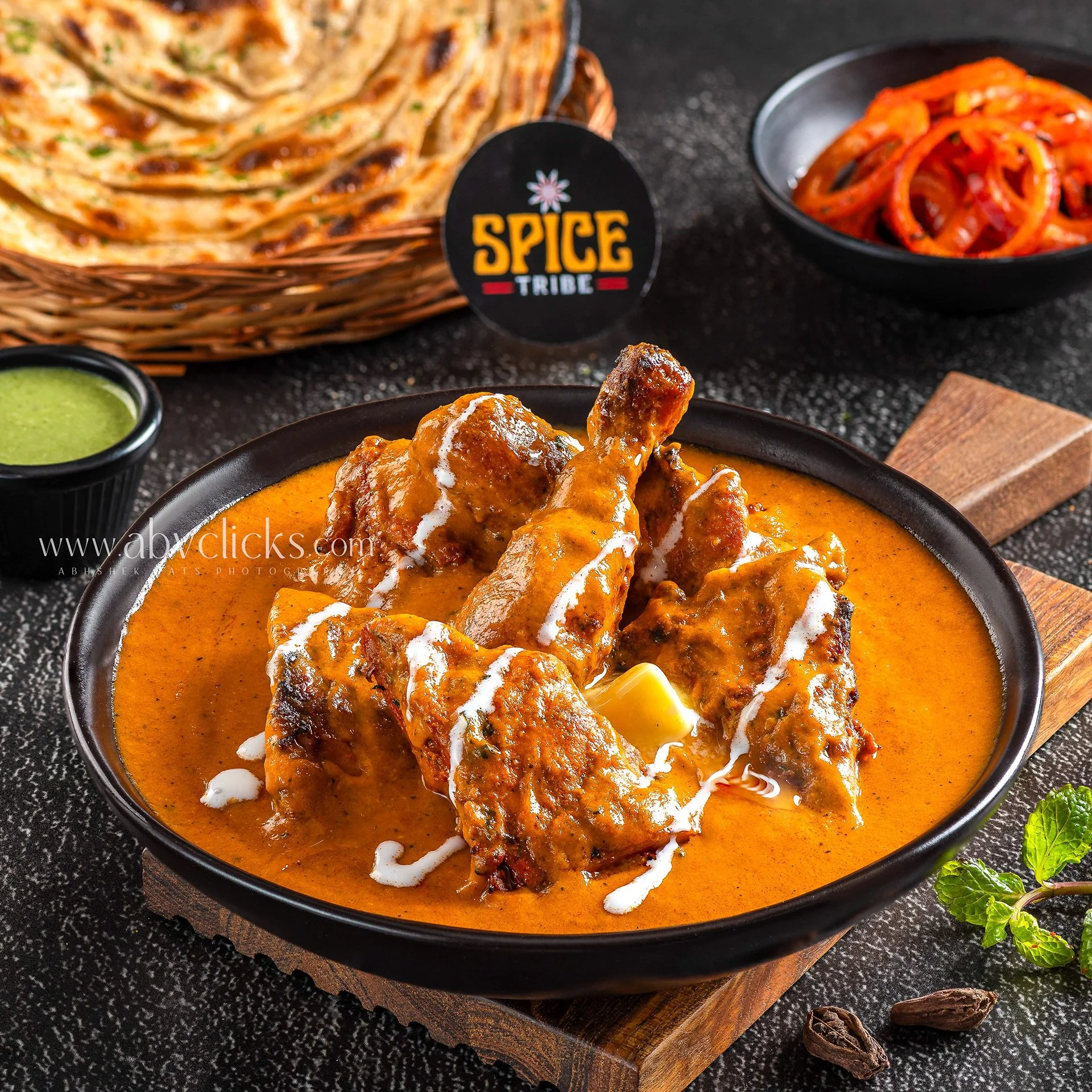 A black bowl of Indian butter chicken curry garnished with cream, placed on a wooden board. To the side, a small black bowl of green chutney, fresh mint leaves, and a lit cinnamon stick are visible. In the background, a basket of Naan bread and a bla