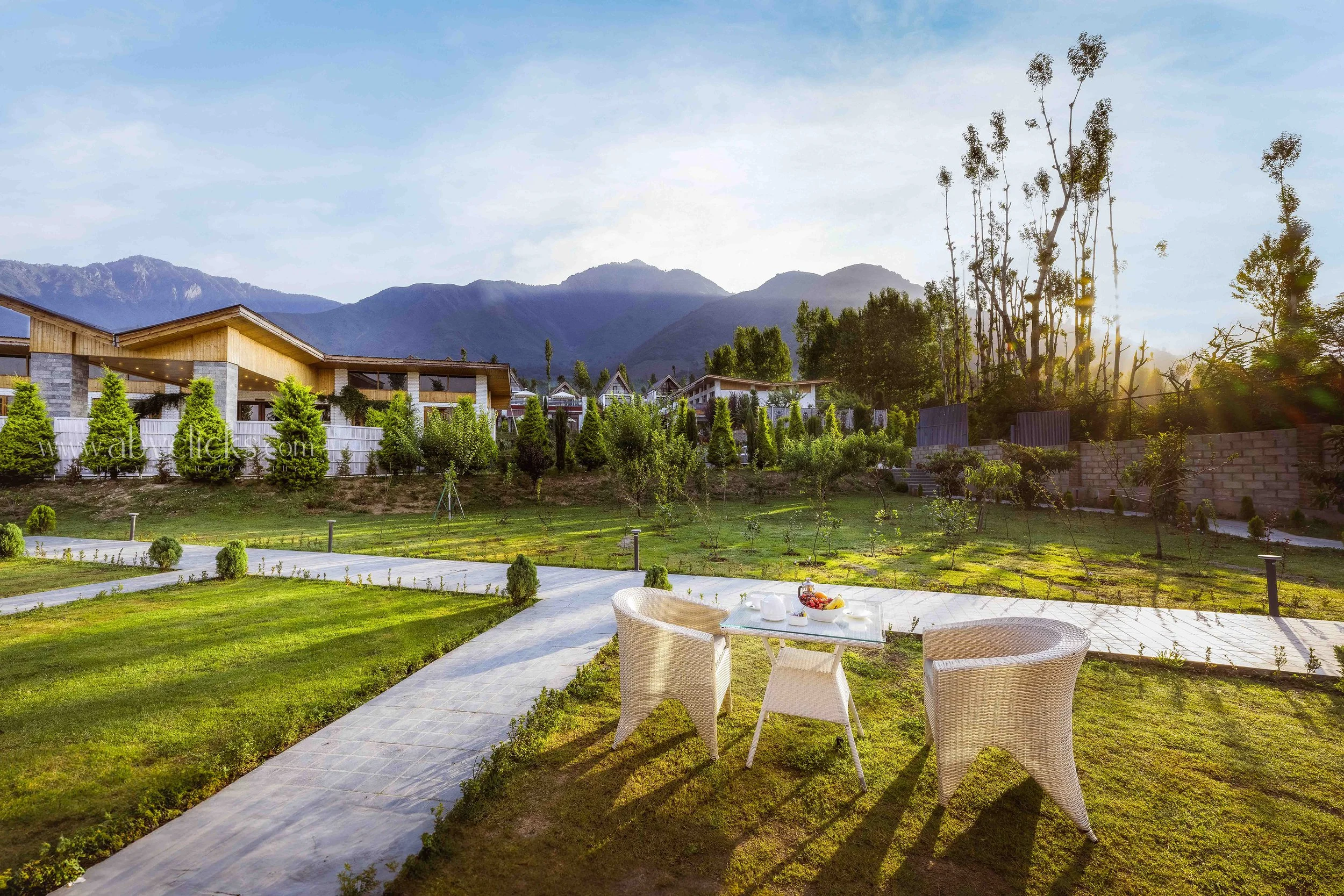 A landscaped outdoor patio area with a table set for a meal, white chairs, lush green grass, trees, modern houses, and mountains in the background during sunset.