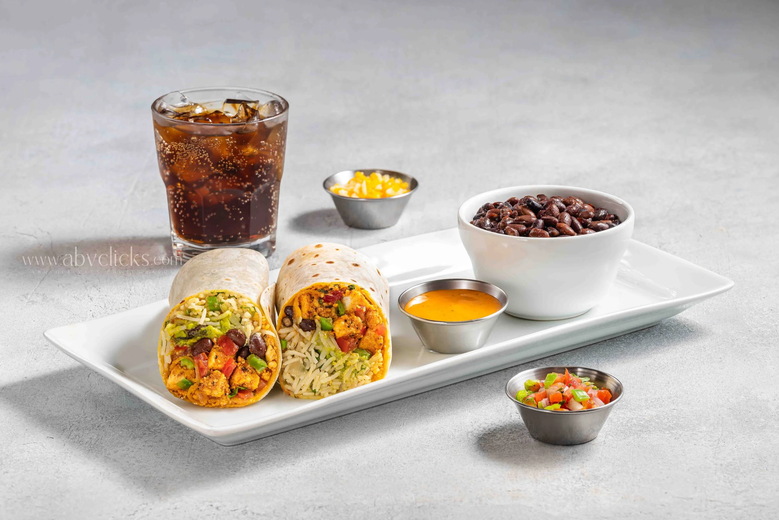 A white rectangular plate with three burritos filled with rice, beans, and vegetables, accompanied by small bowls of salsa and sauce, a bowl of black beans, a small bowl of shredded cheese, a glass of iced cola, and some condiments, all on a light gr