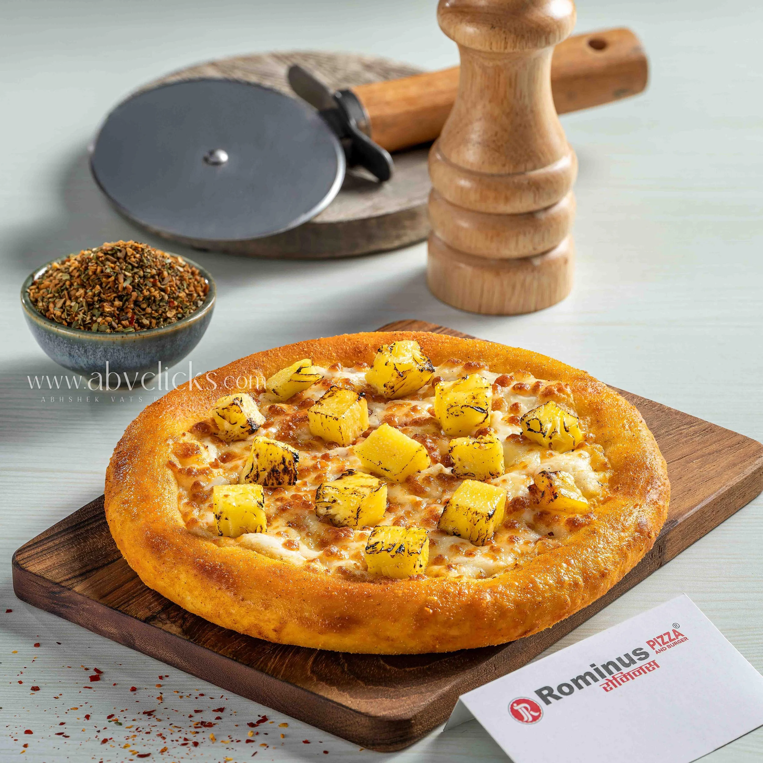 A cheese pizza with grilled pineapple pieces on top, placed on a round wooden cutting board. In the background, there is a bowl of crushed red pepper flakes, a pizza cutter, and a wooden rolling pin. There is a Domino's Pizza box partially visible be
