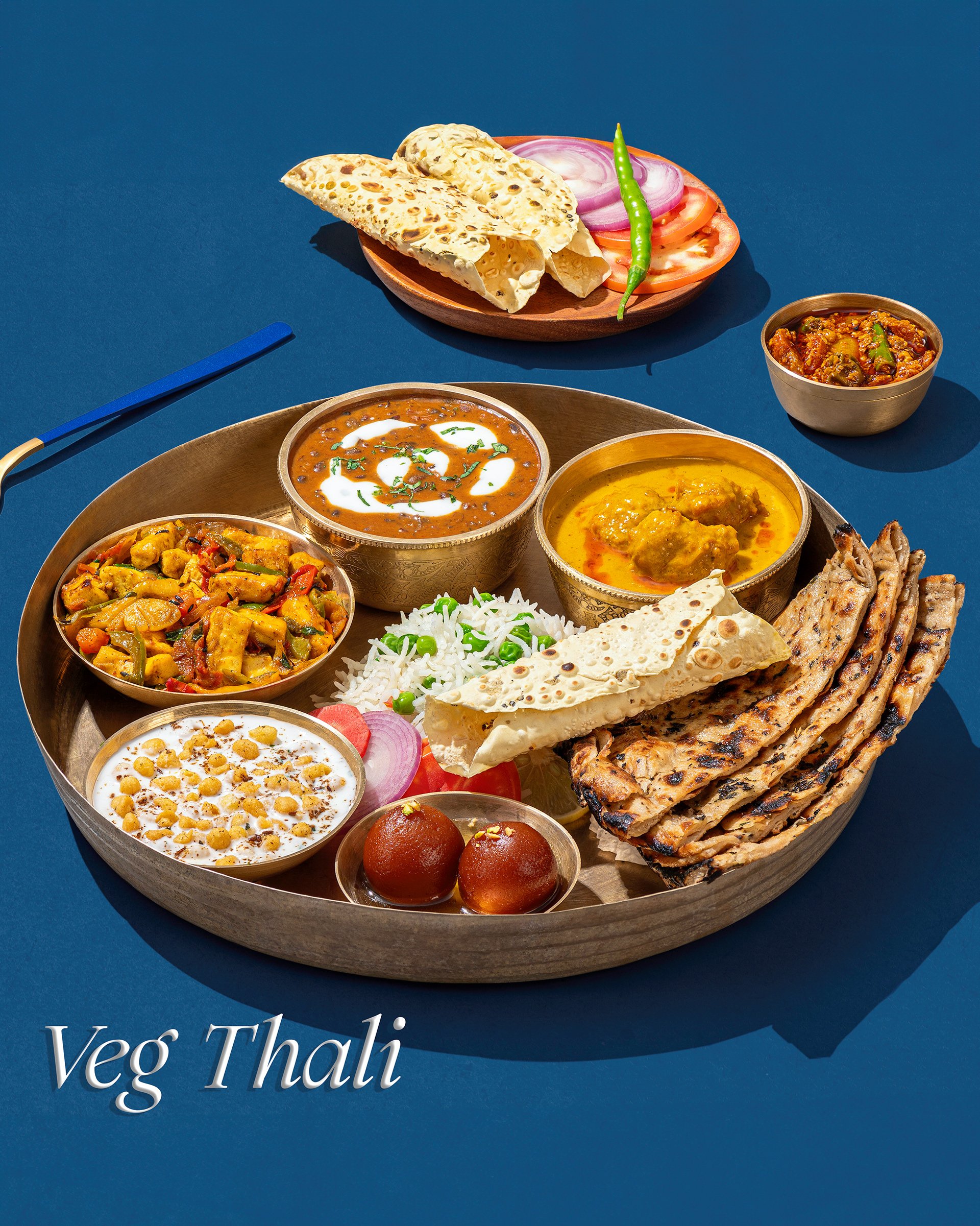 A traditional vegetarian Indian Thali with assorted dishes, including curries, rice, naan, and sweets, served on a round wooden tray. There is also a side plate with sliced vegetables, a green chili, tomato slices, onion slices, a bowl of spicy curry