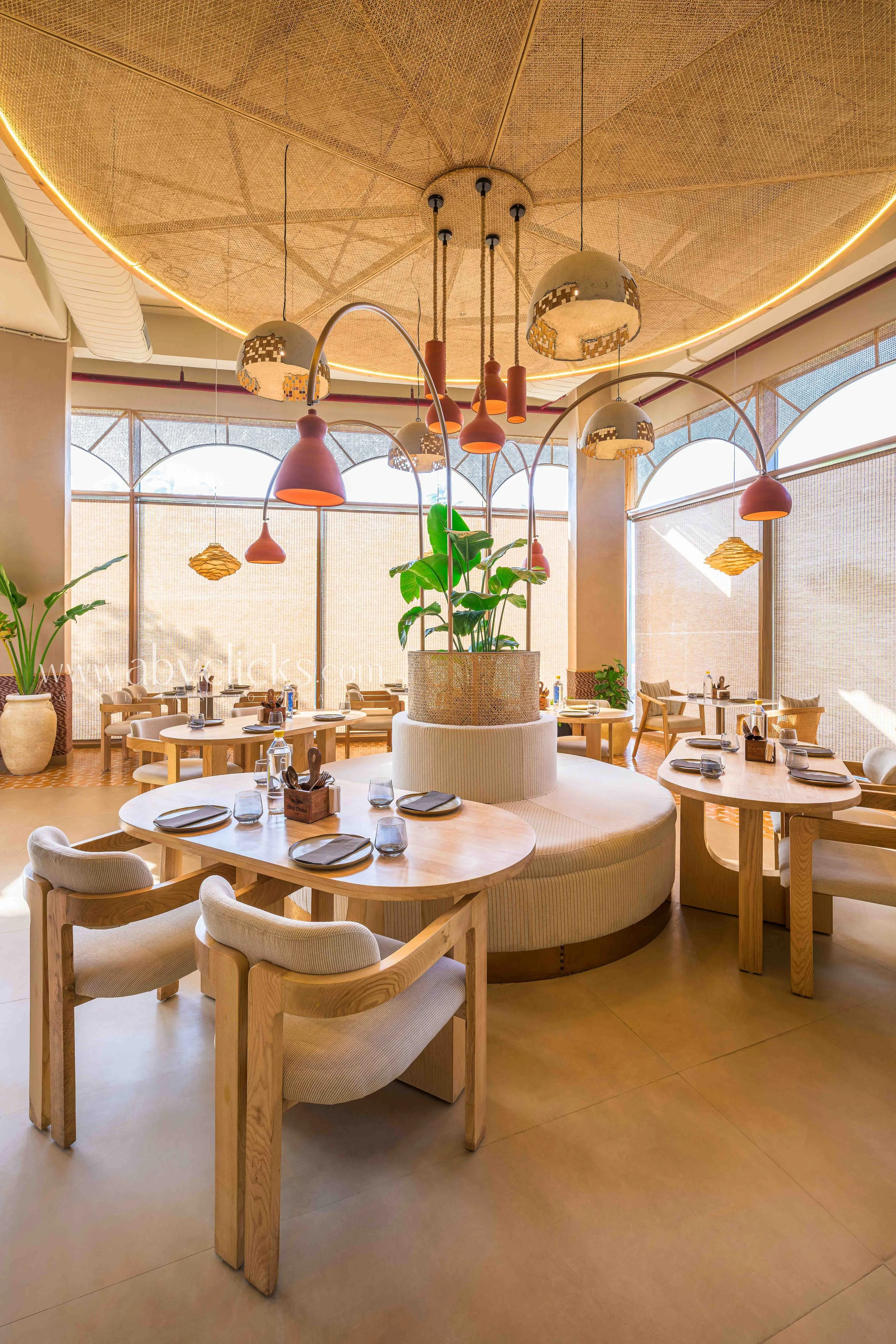 Contemporary restaurant interior with round tables, beige chairs, and a large circular central seating area with a tall plant. Modern ceiling lights and pendant lamps hang from the ceiling, which has inlaid wooden paneling and built-in lighting. Larg