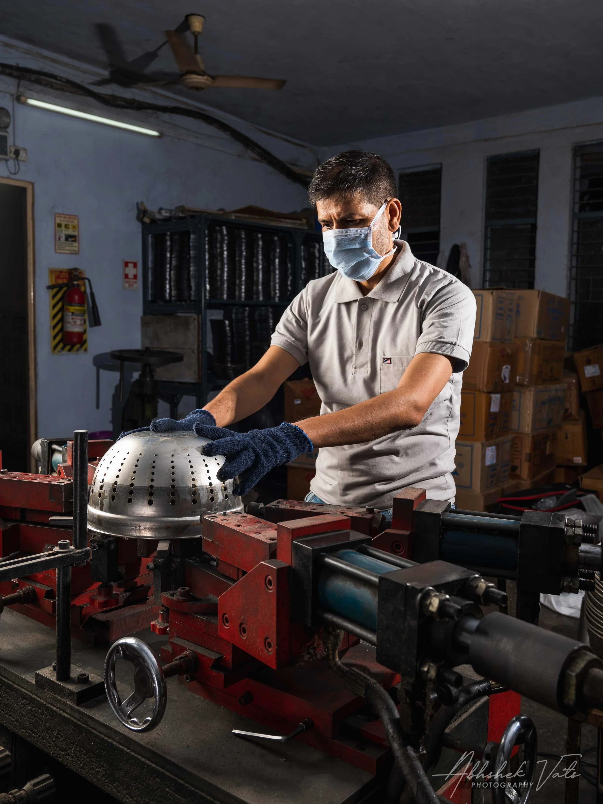 A man wearing a gray polo shirt and a face mask working with machinery in an industrial setting, surrounded by boxes and equipment.