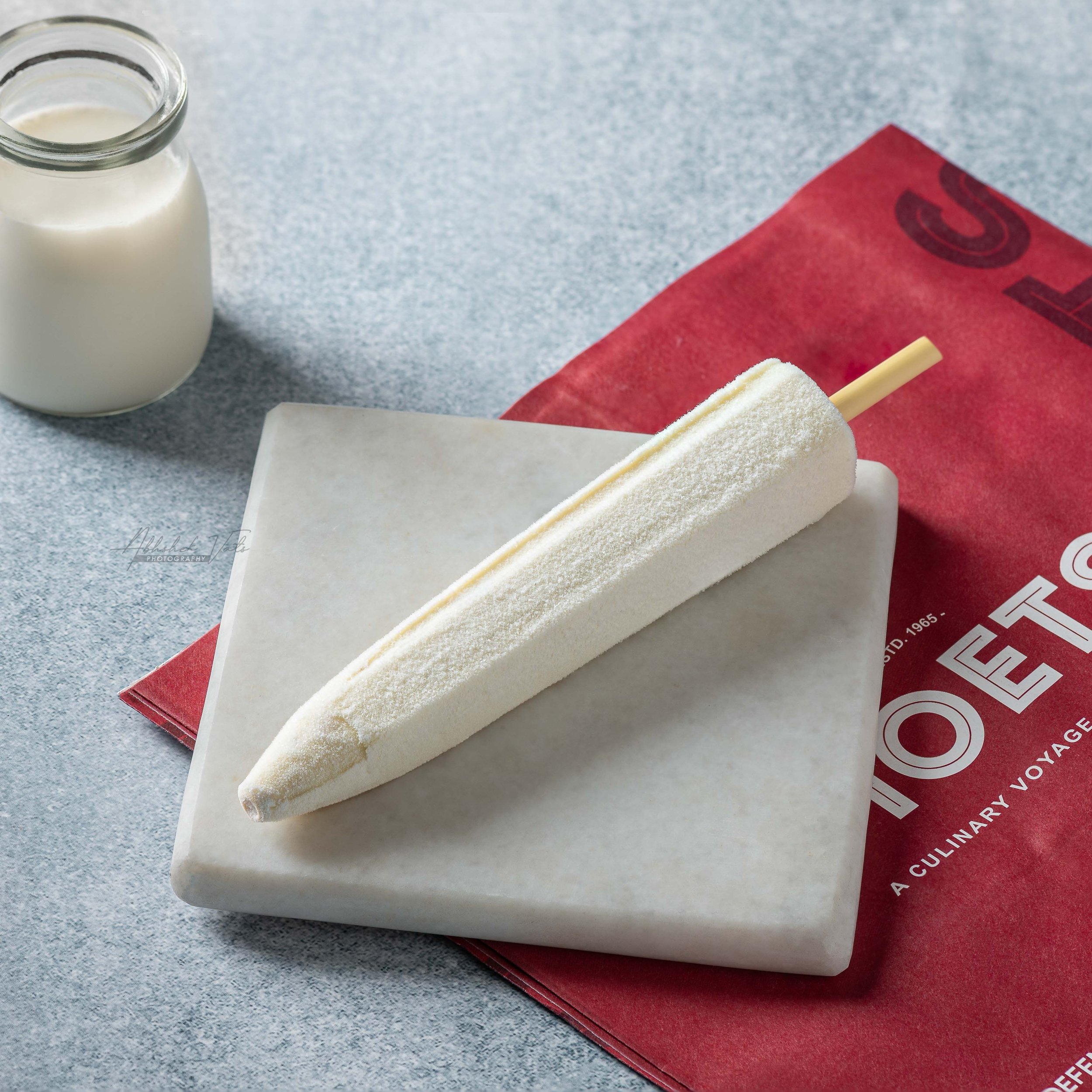 A bottle of milk, a red menu or book, and a white cake with a yellow candle on a gray surface.