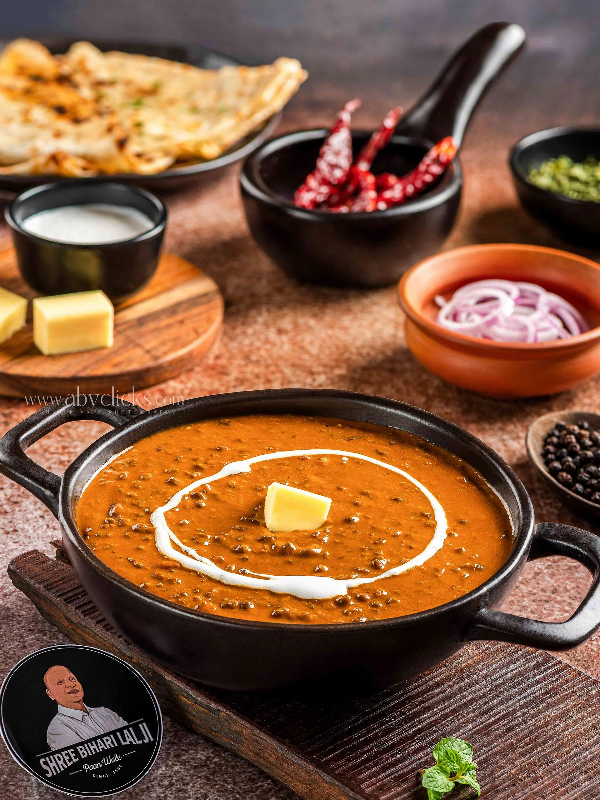 A pot of Indian lentil curry garnished with a swirl of cream and a piece of butter, surrounded by bowls of chopped onions, dried red chilies, chopped herbs, and black peppercorns on a rustic wooden surface.