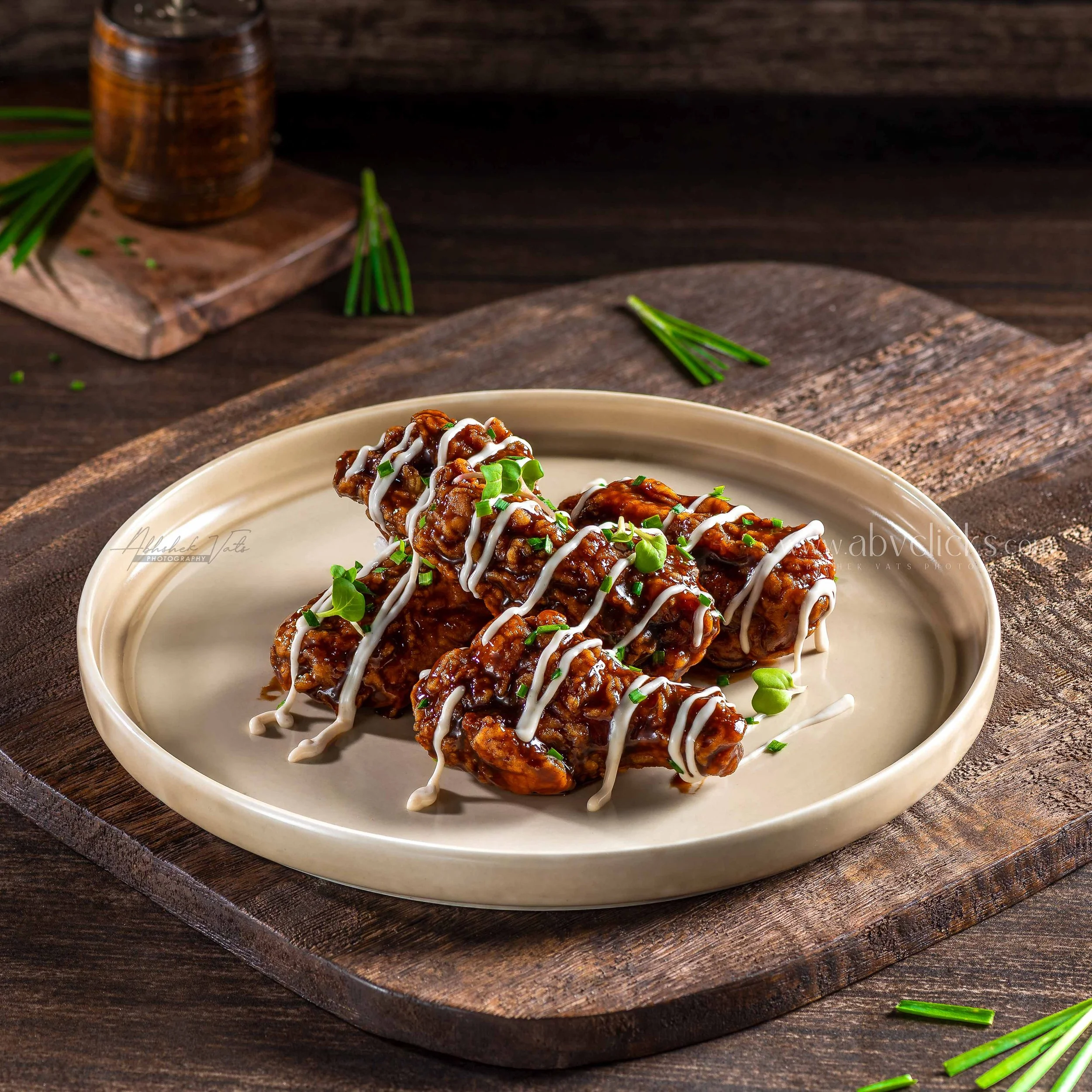 Close-up of a plate of Asian-style chicken drumettes coated in sauce, topped with chopped green onions and white sauce drizzle, on a rustic wooden serving board.