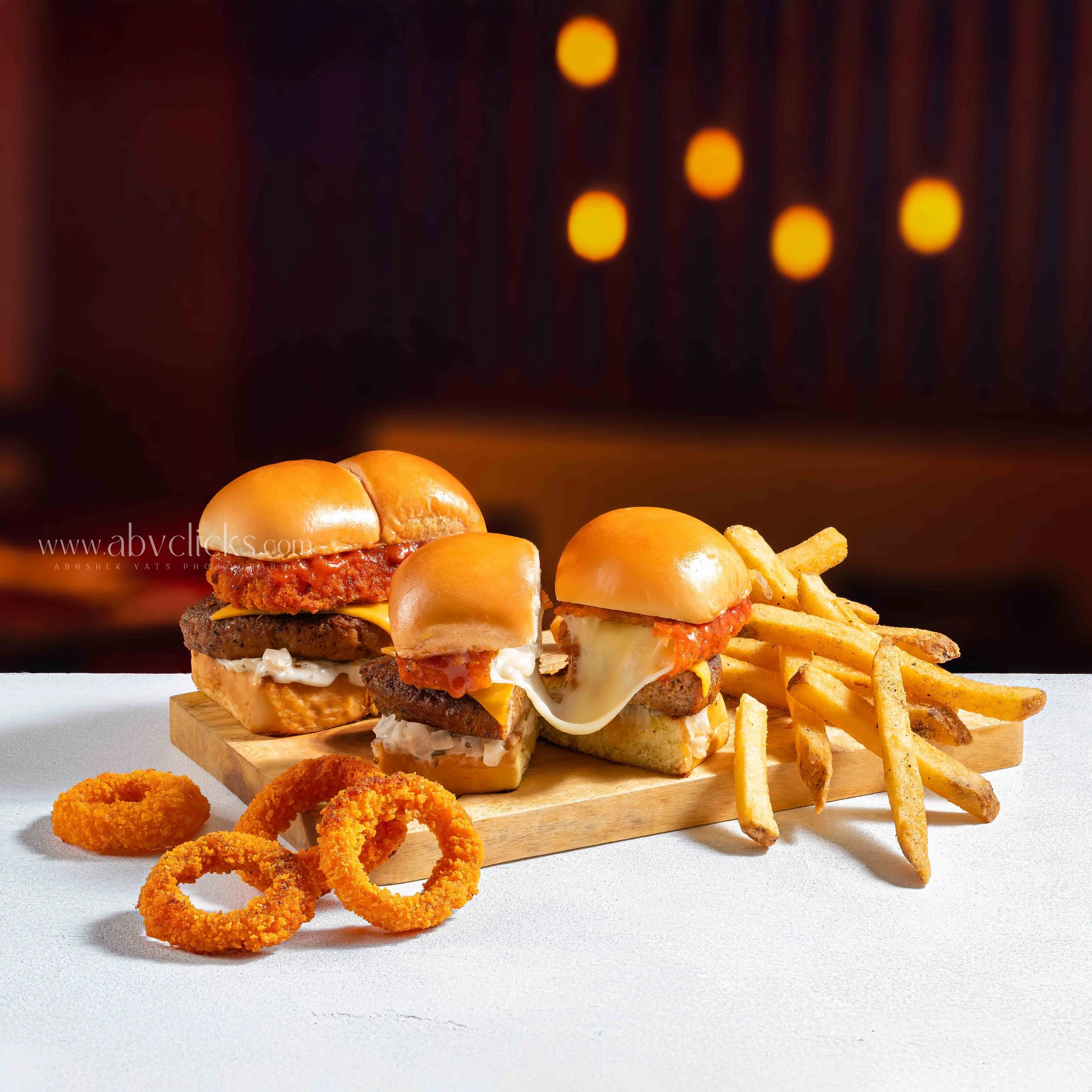 Close-up of cheeseburgers with buns, cheese, and meat patties, served on a wooden board, with fried onion rings and potato chips on a light-colored surface.