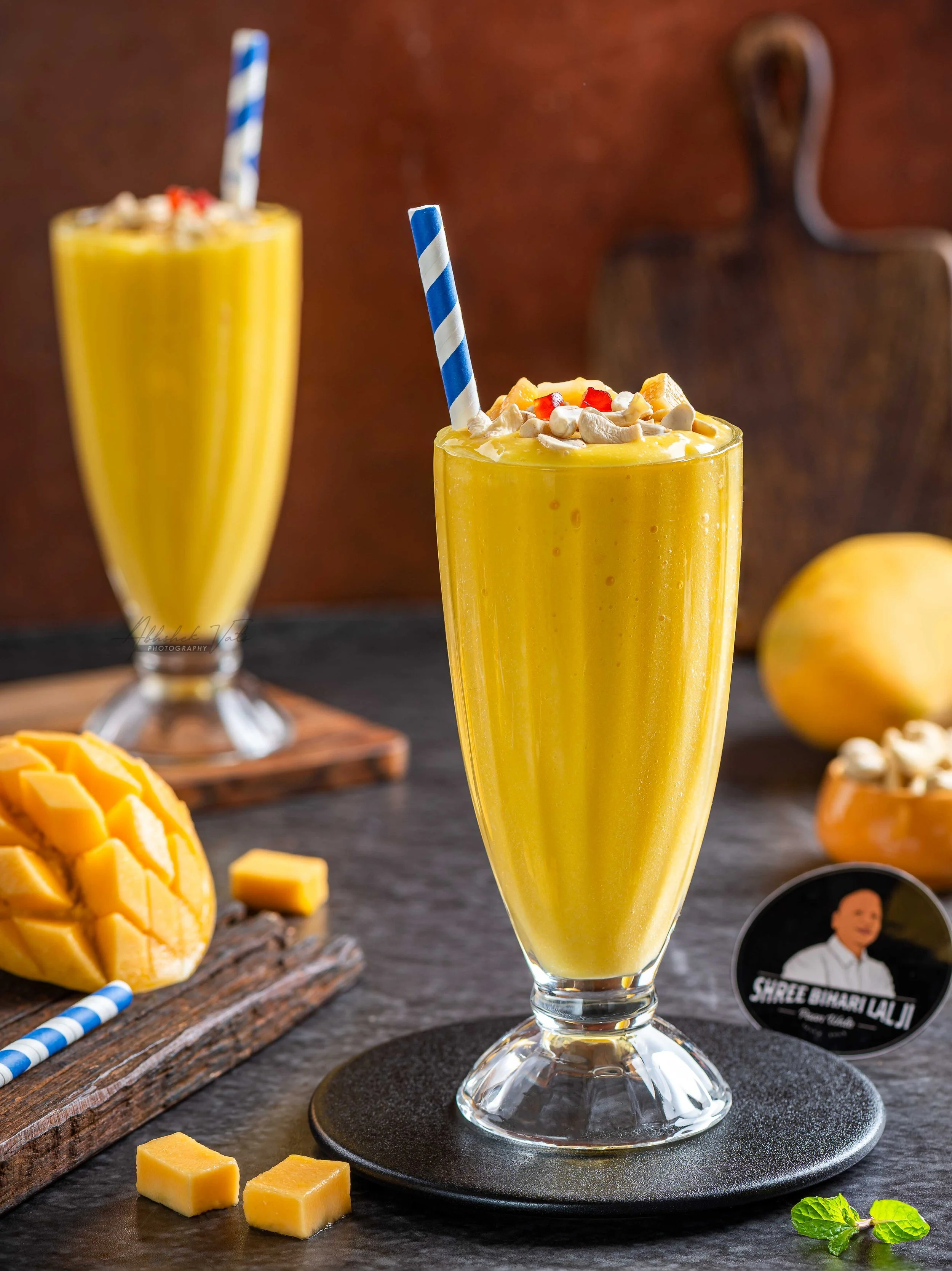 Tall glass of mango smoothie topped with cashews, pineapple chunks, and pomegranate seeds, with a blue and white striped straw, on a black coaster, with a mango and mint leaves nearby, and a second similar smoothie in the background.
