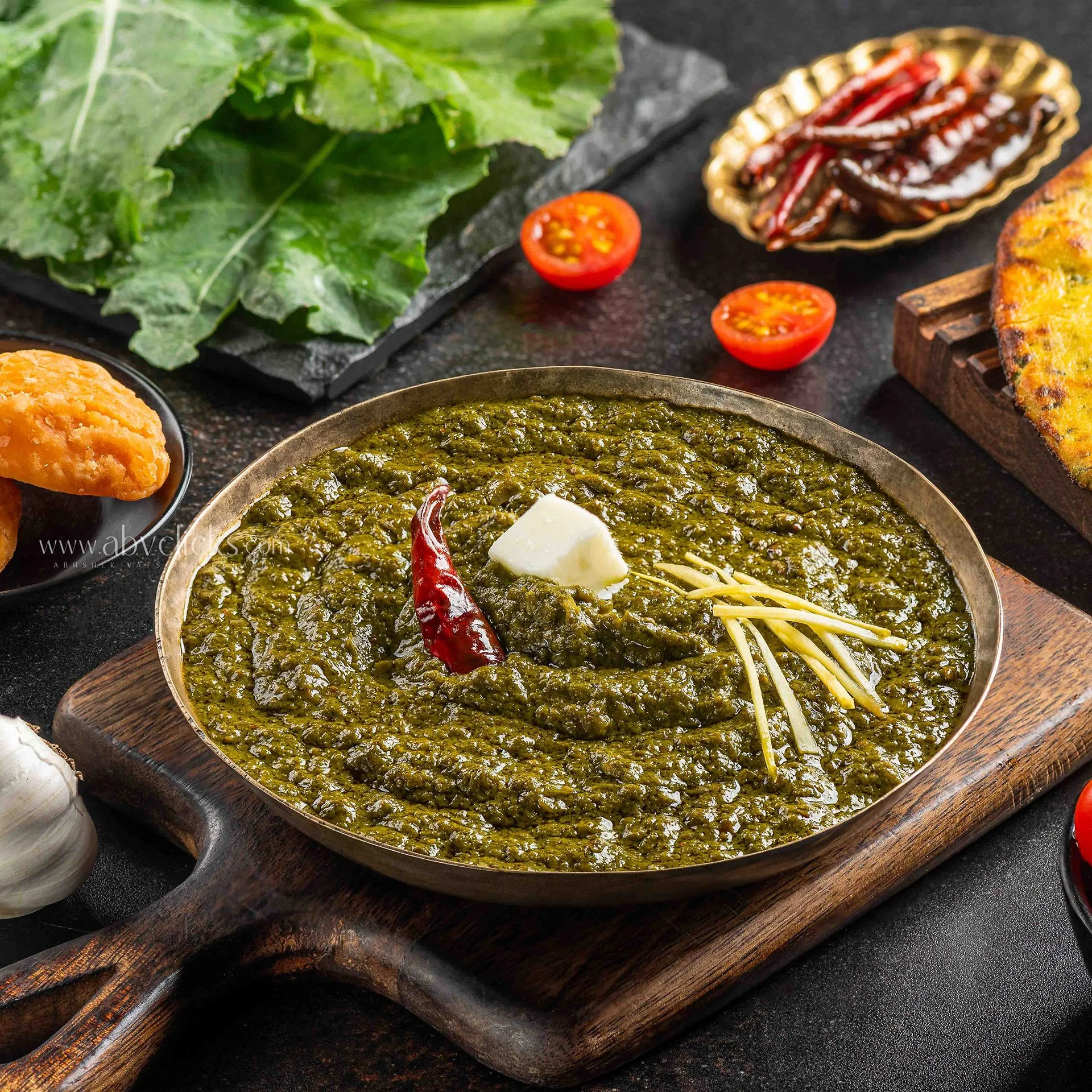 A bowl of cooked green spinach with a dollop of butter, set on a wooden cutting board. Around it, there are cherry tomatoes, a plate of dried red chilies, and a piece of grilled bread with herbs. The background includes fresh leafy greens and a black