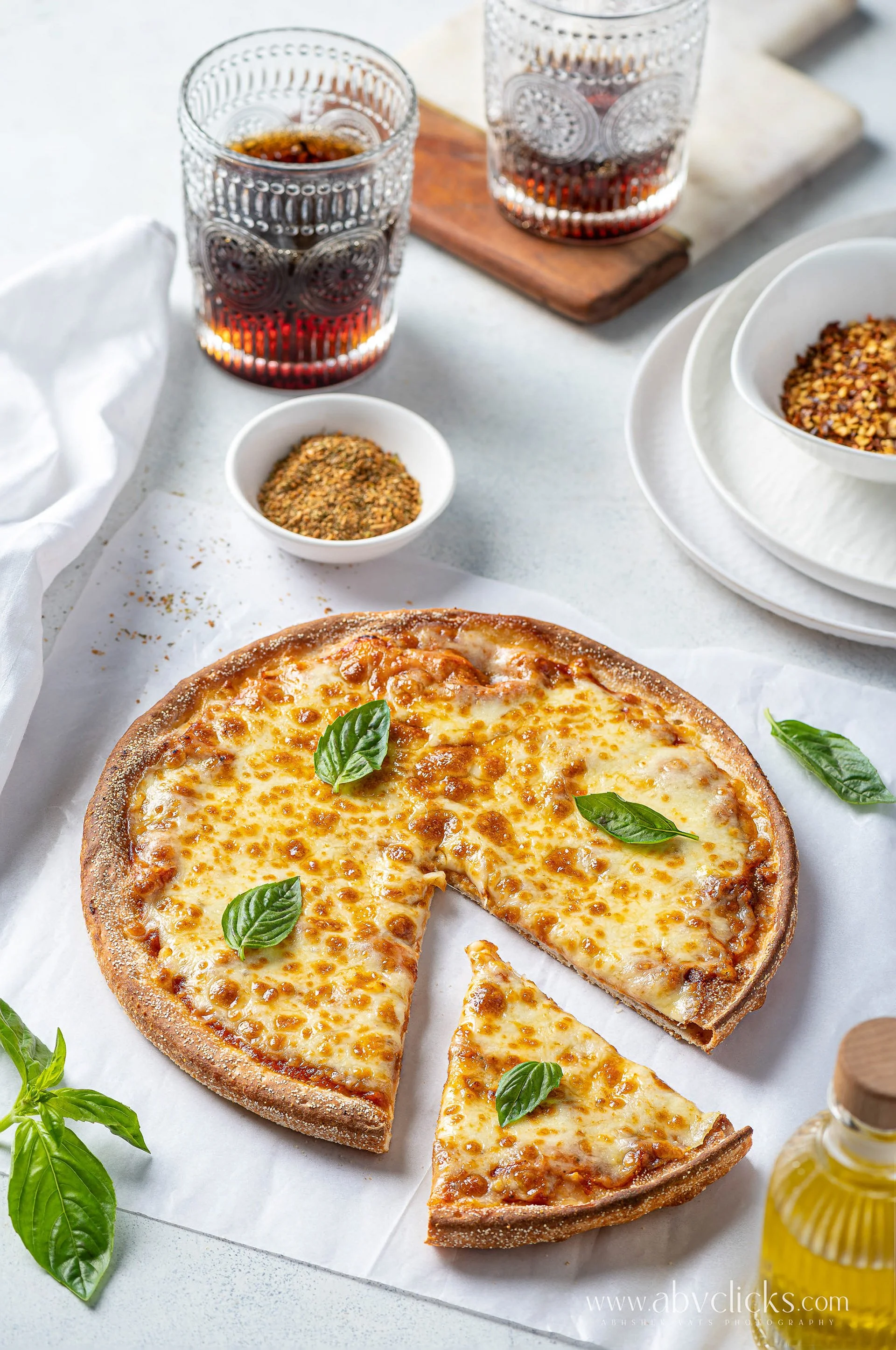 Cheese pizza on white parchment paper with fresh basil leaves, two glasses of soda, small bowl of crushed red pepper, and olive oil bottle on white table.