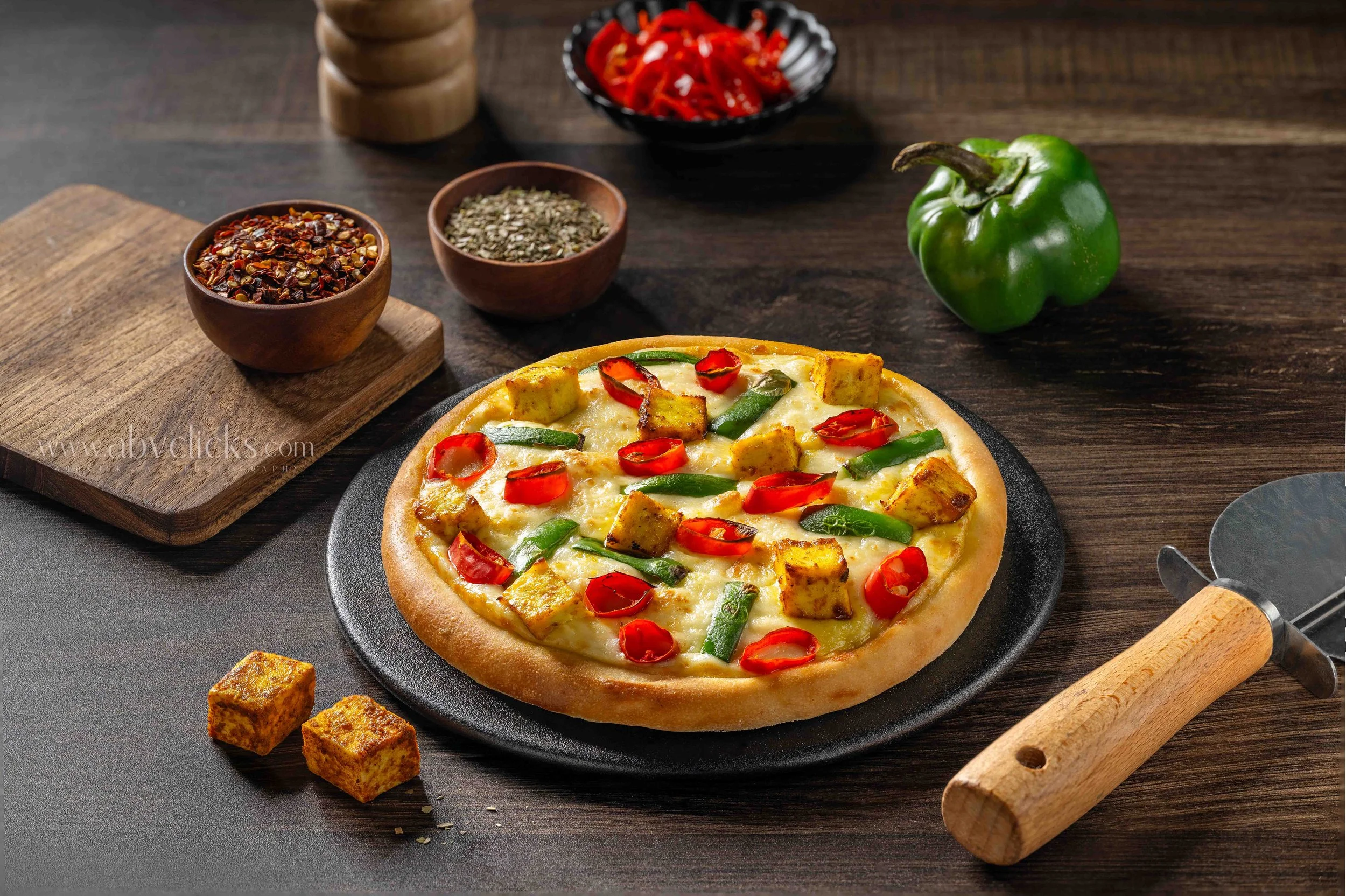 A freshly baked pizza topped with cheese, cherry tomatoes, green peppers, and seasoned tofu chunks on a black slate plate, with bowls of crushed chili peppers and herbs, a red bell pepper, a wooden pepper grinder, and a rolling pin on a dark wooden t