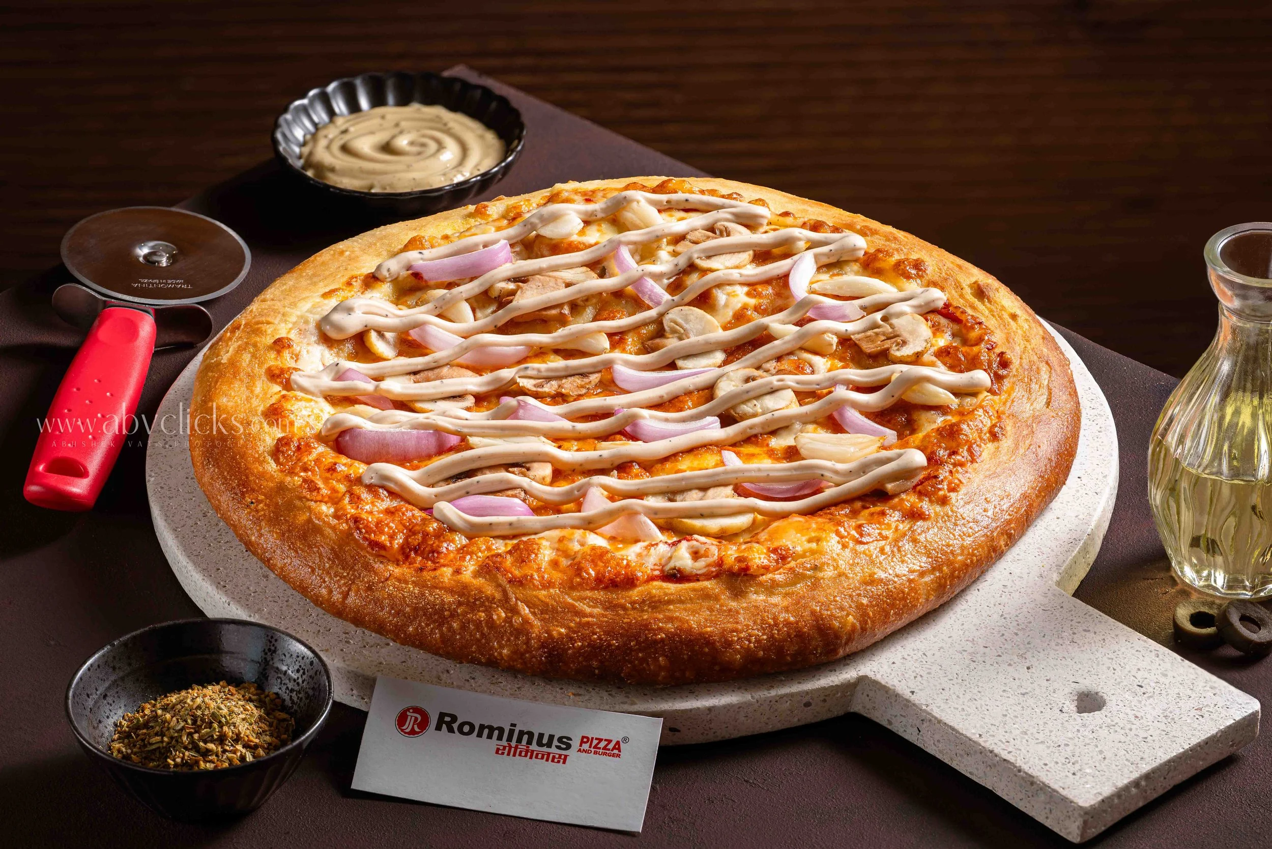 A pizza with a thick crust topped with chicken, red onions, and drizzled with white sauce. It is served on a white platter with small bowls of dipping sauces and spices nearby.
