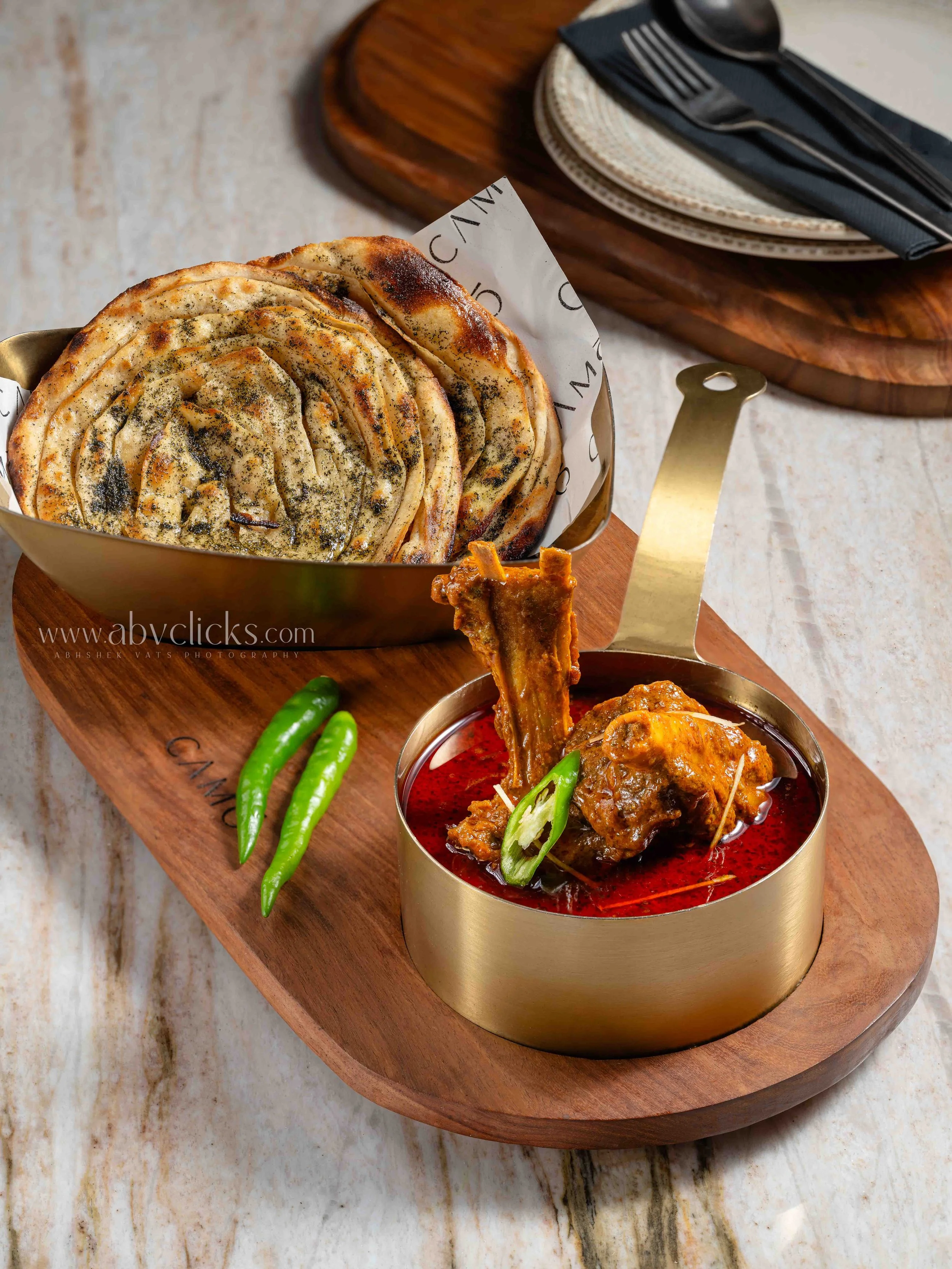 A bowl of grilled spiral bread, a bowl of spicy meat curry, and two green chilies on a wooden serving board.