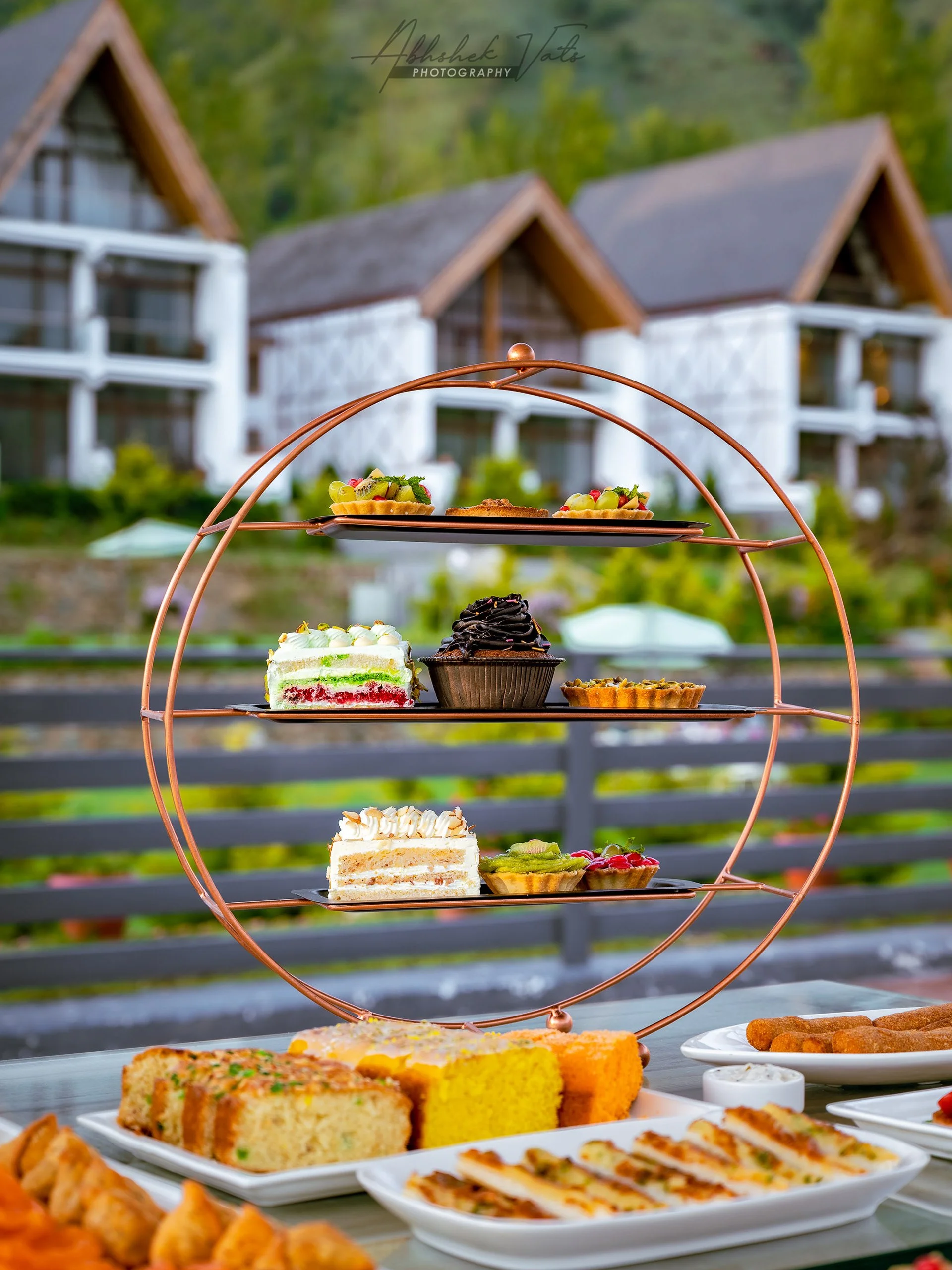 A tiered metal stand displaying assorted cakes and desserts outdoors with a blurred background of houses and greenery.