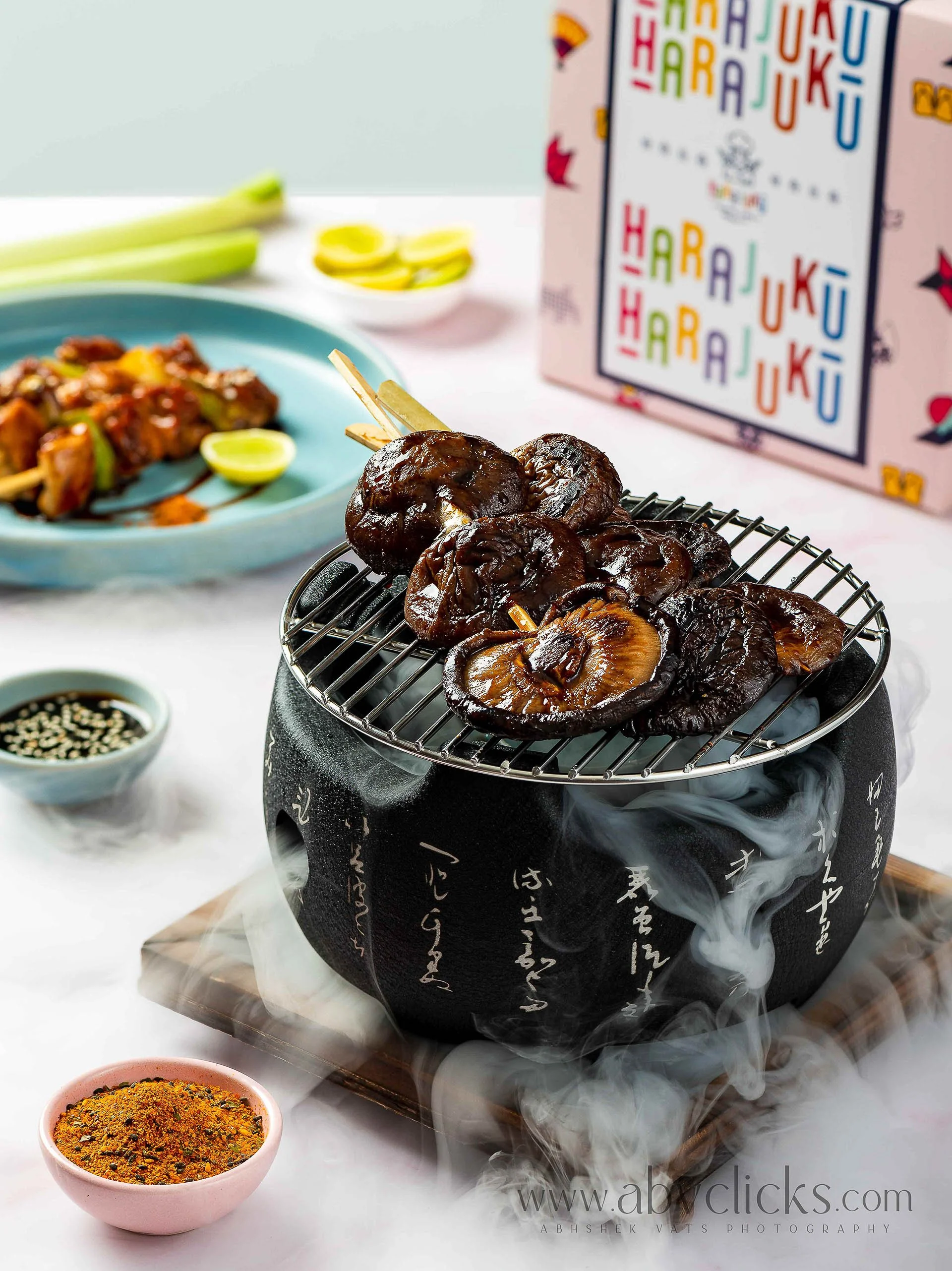Smoked mushrooms on a traditional Japanese charcoal grill with smoke rising, surrounded by small bowls of spices and sauces, with a box of colorful greeting cards in the background.