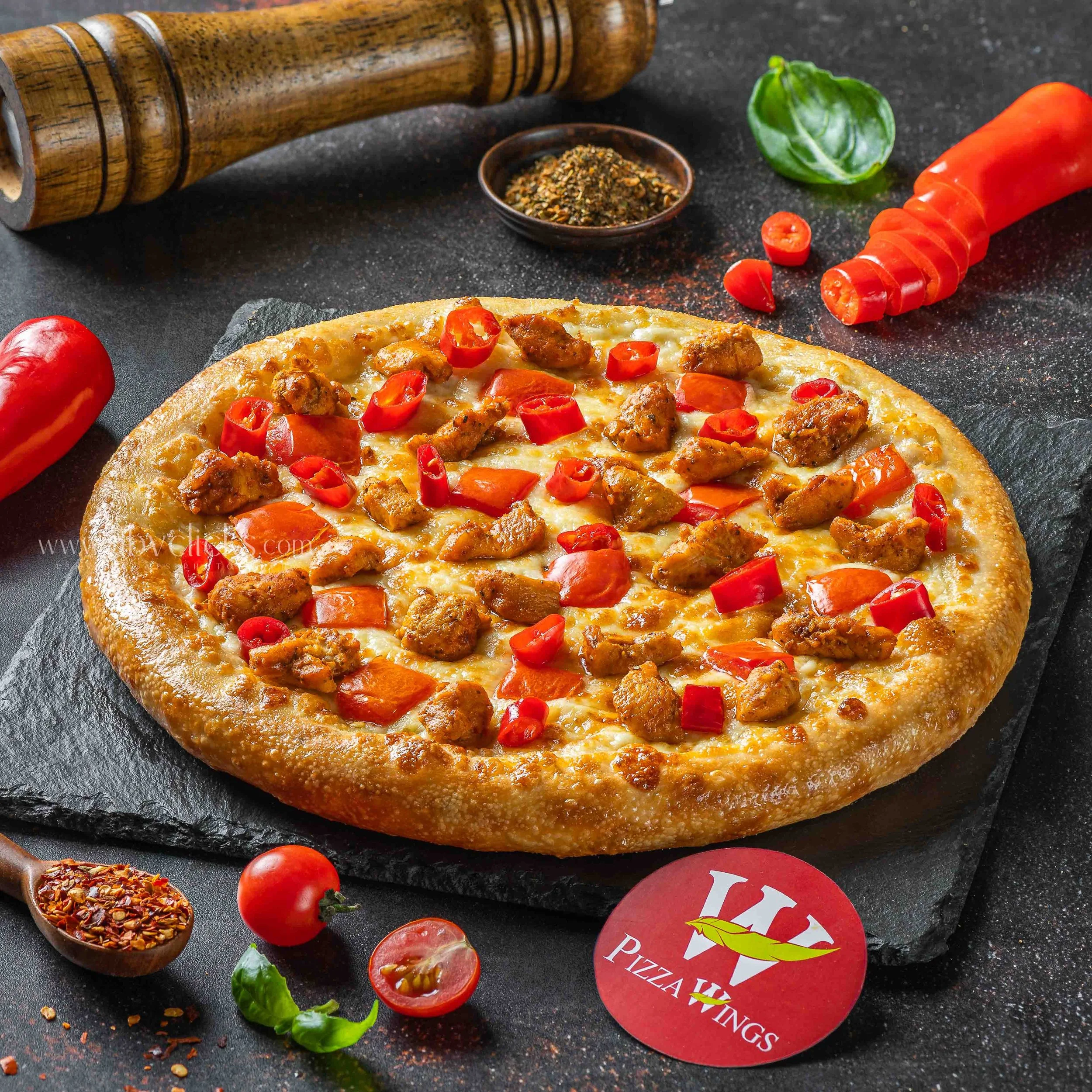 A freshly baked pizza with melted cheese, sliced red chili peppers, and cooked ground meat, on a black surface surrounded by cherry tomatoes, basil leaves, and a red circular pizza restaurant sign with the words 'Pizza' and 'Tings' visible.