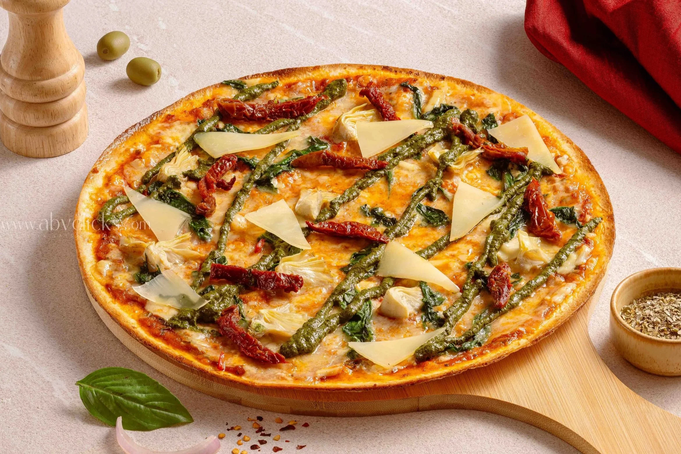 A pizza with cheese, green pesto, sun-dried tomatoes, artichoke hearts, and basil leaves on a wooden board, surrounded by small bowls of spices and a basil leaf.