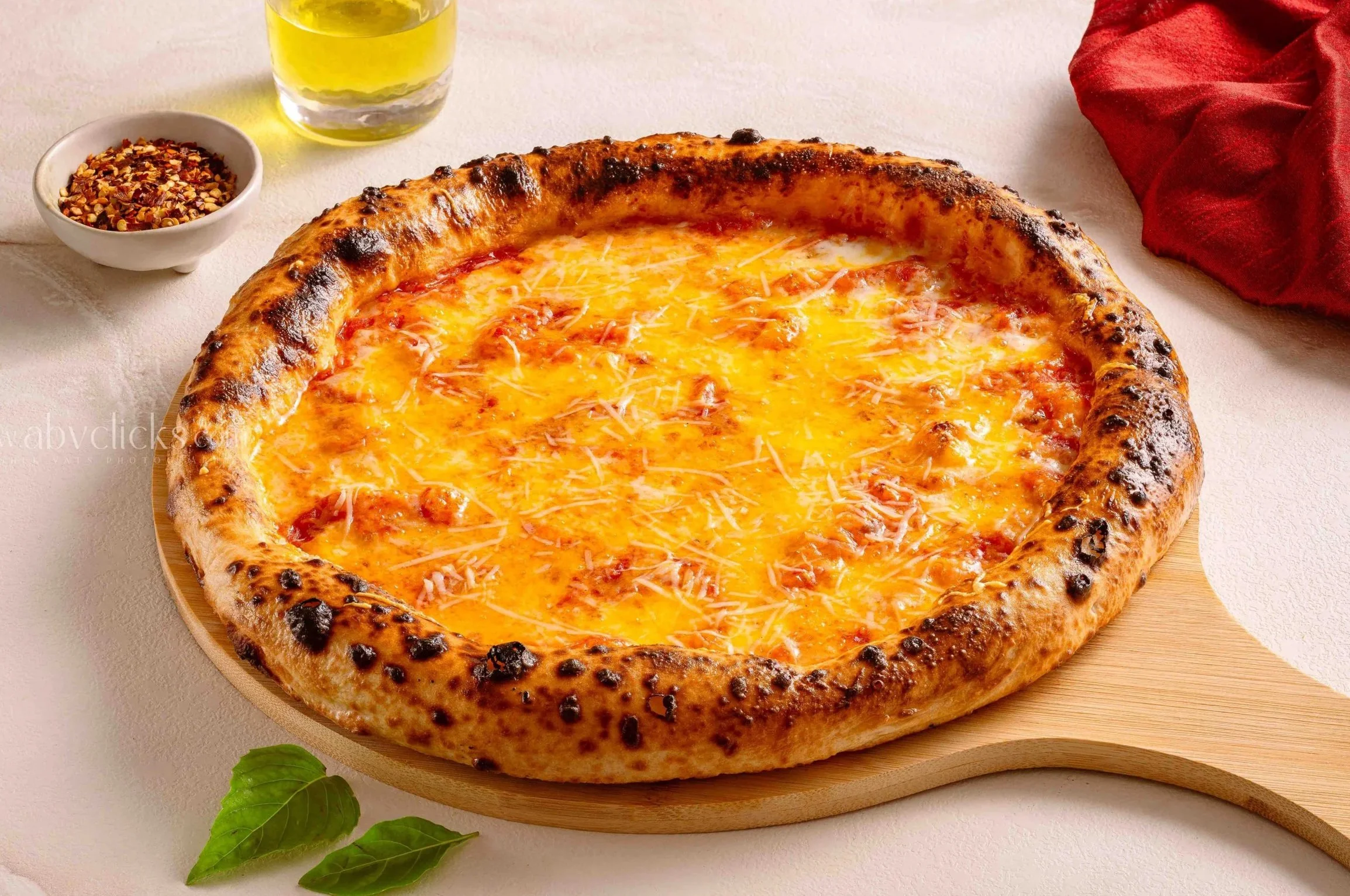 Cheese pizza on a wooden board with a glass of yellow drink, a small bowl of red pepper flakes, and fresh green basil leaves.