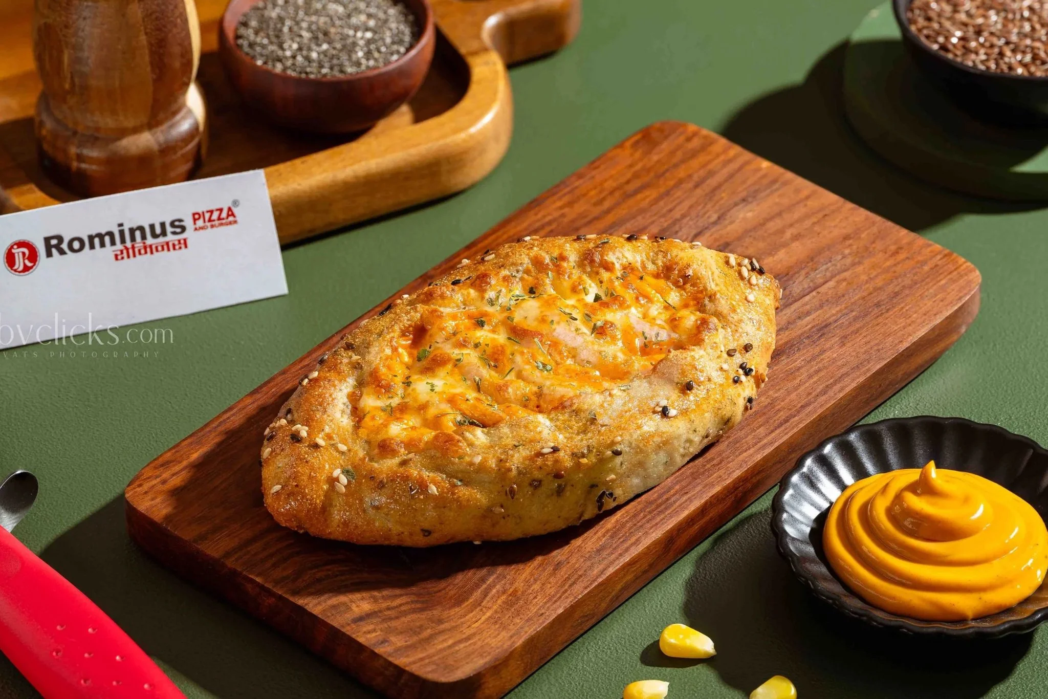 A piece of garlic bread topped with cheese and herbs on a wooden serving board, with small bowls of mustard and black sesame seeds, and a label reading 'Rominus Pizza and Burger' in the background.