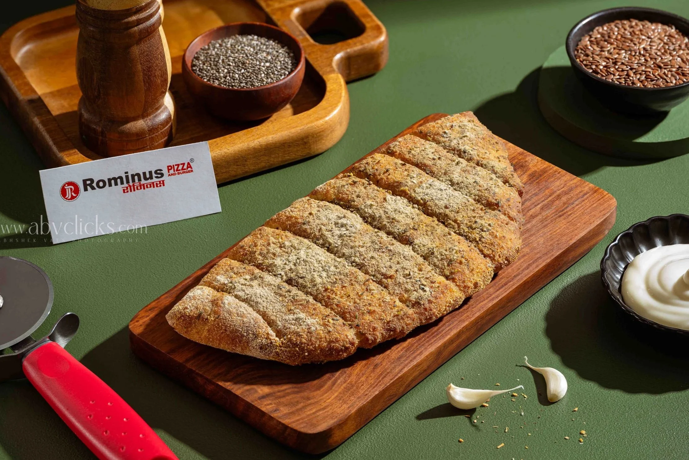 A wooden cutting board with a crusted baked bread or flatbread sliced into pieces, with scattered garlic cloves and a small dish of mayonnaise beside it. In the background, a black bowl filled with flax seeds, a wooden tray with small bowls of black 