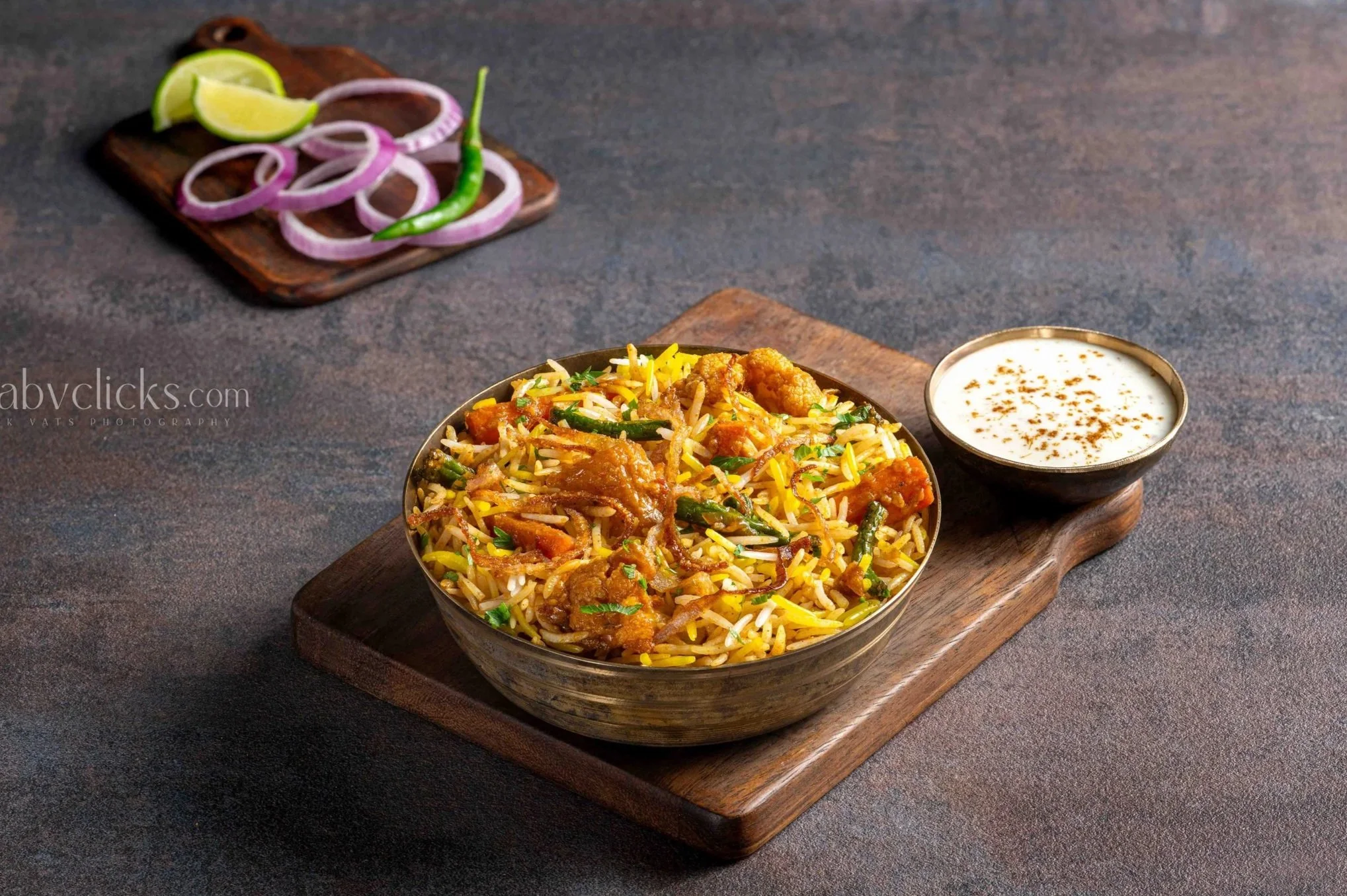 A bowl of Indian chicken biryani on a wooden tray with a bowl of raita on the side, a plate of sliced onions and lime wedges in the background.