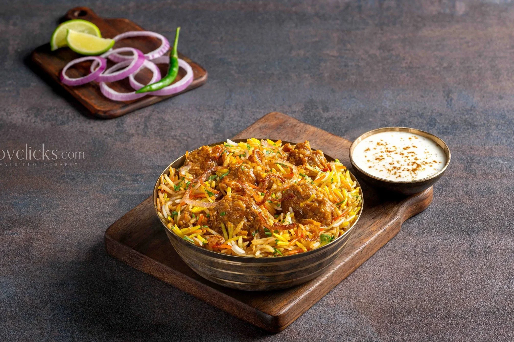 A bowl of colorful Indian chicken biryani with herbs and spices, a small bowl of raita, and a plate of sliced red onions, green chili, and lime wedges on a wooden board on a dark surface.
