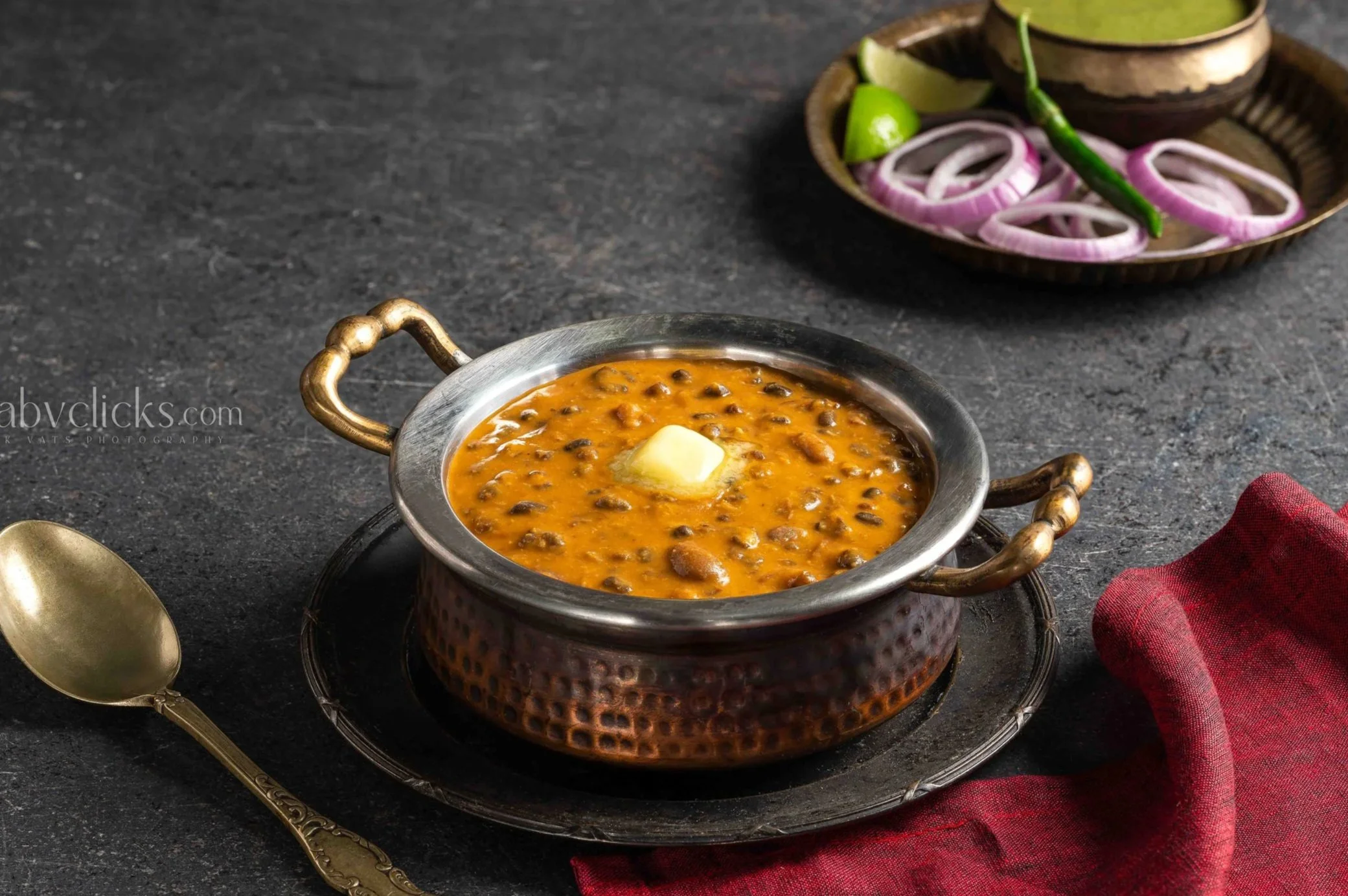 A traditional Indian dish called dal served in a copper bowl with a pat of butter on top, accompanied by sliced red onions, lime wedges, and green chili on a dark textured surface.