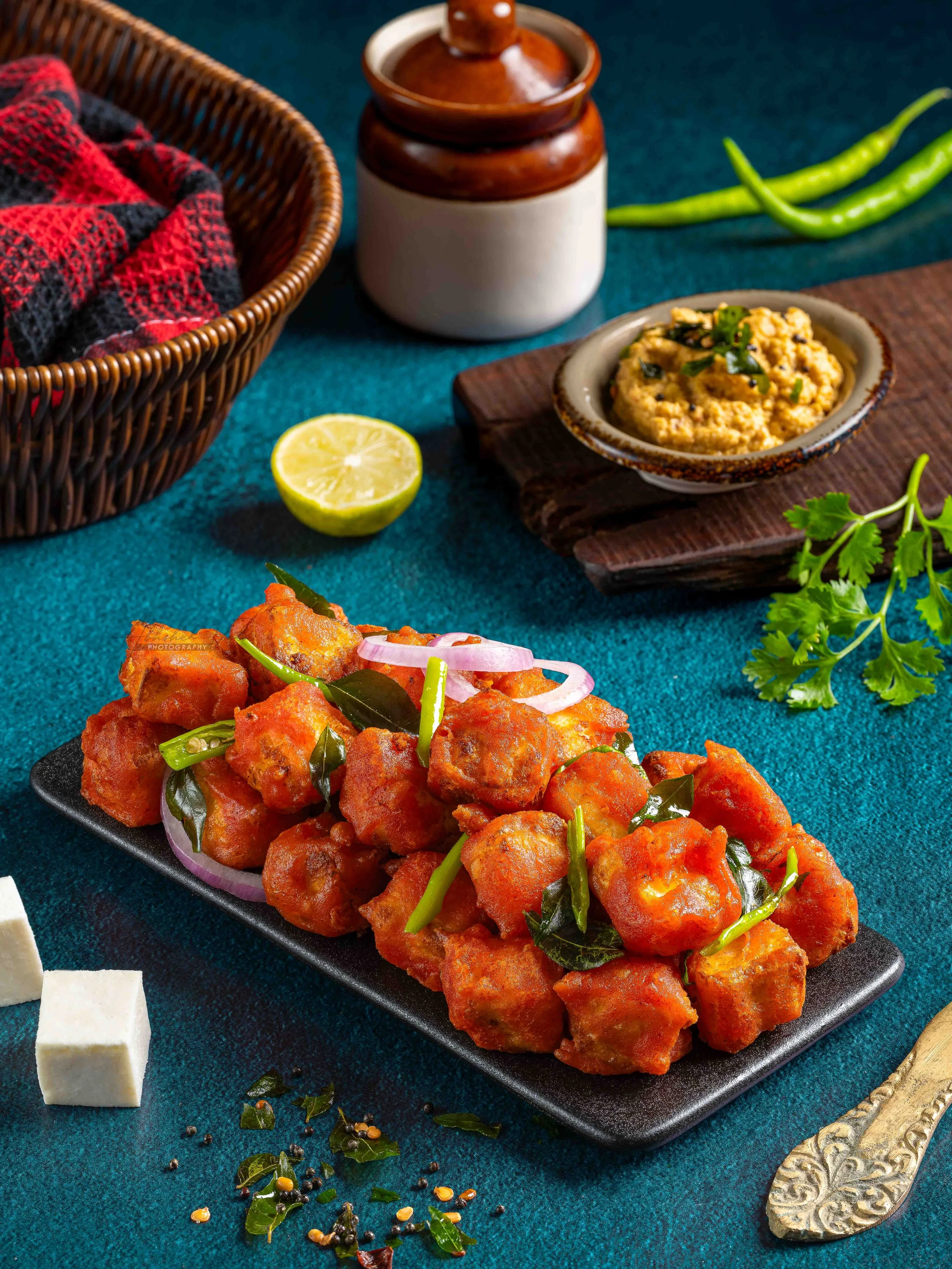 Plate of spicy Indian chicken curry garnished with green chilies and curry leaves, on a dark teal table with a carving knife, a lime half, and scattered seasonings.
