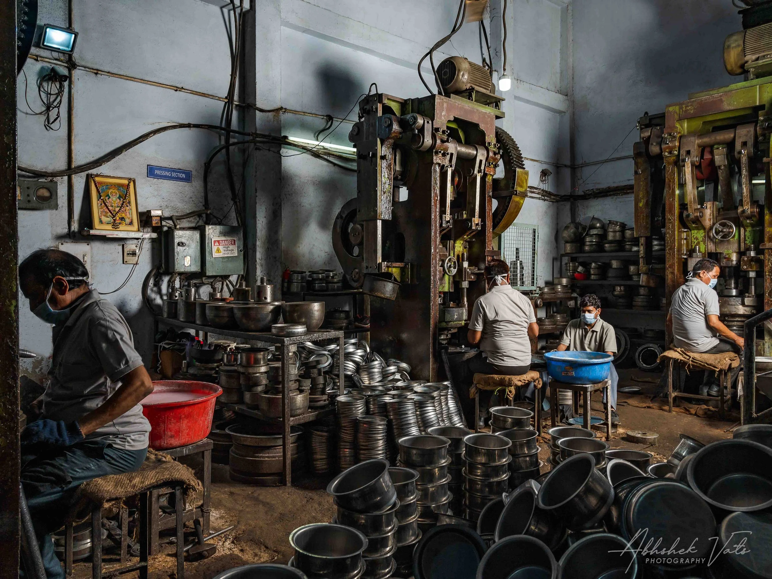 Workers in a factory or workshop surrounded by metal parts, possibly manufacturing or assembling metal containers or components. The workspace appears industrial with machines and tools, and several workers wearing masks.