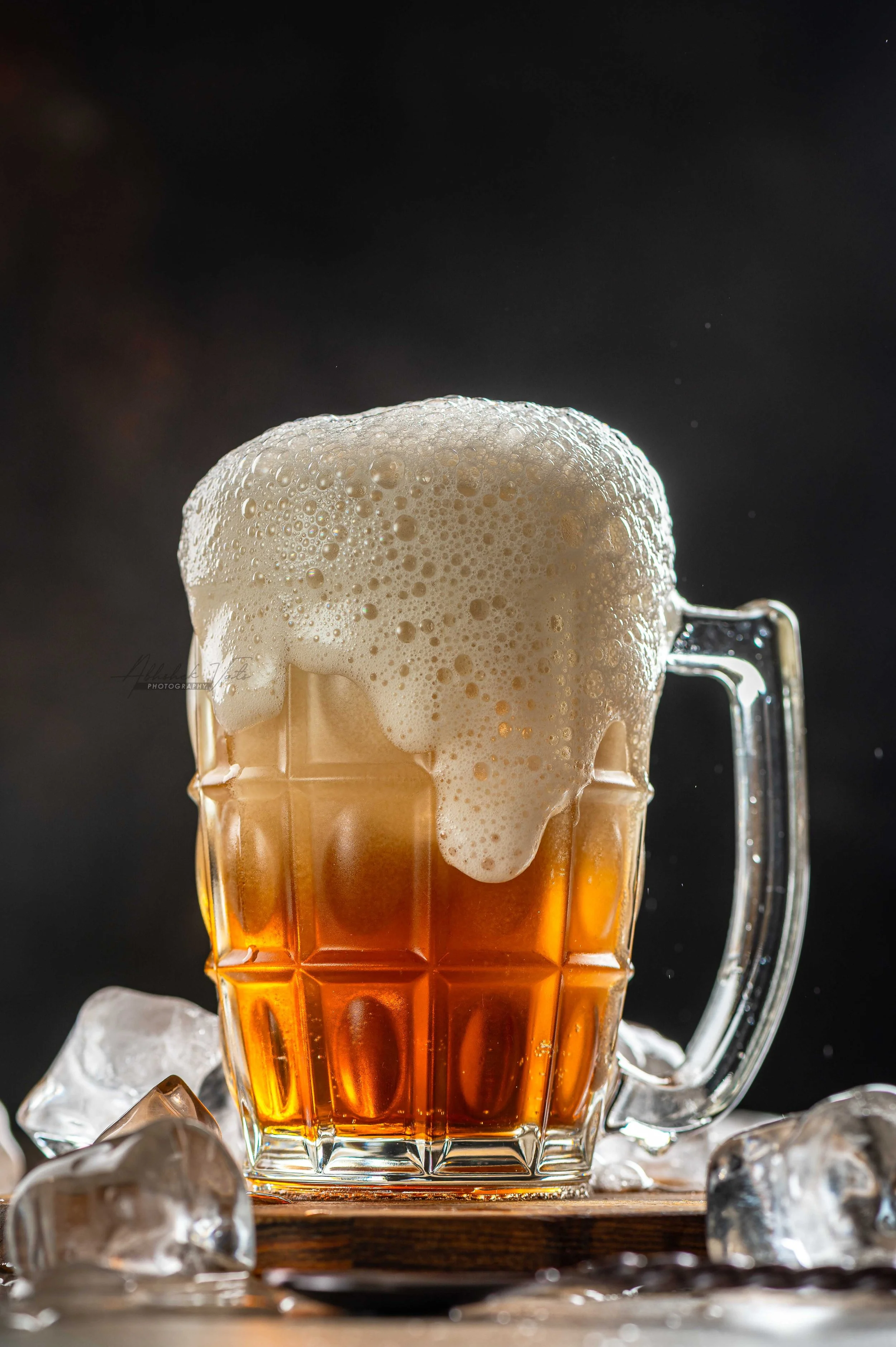 A glass beer mug filled with amber beer and topped with foam, surrounded by ice cubes on a wooden surface against a dark background.