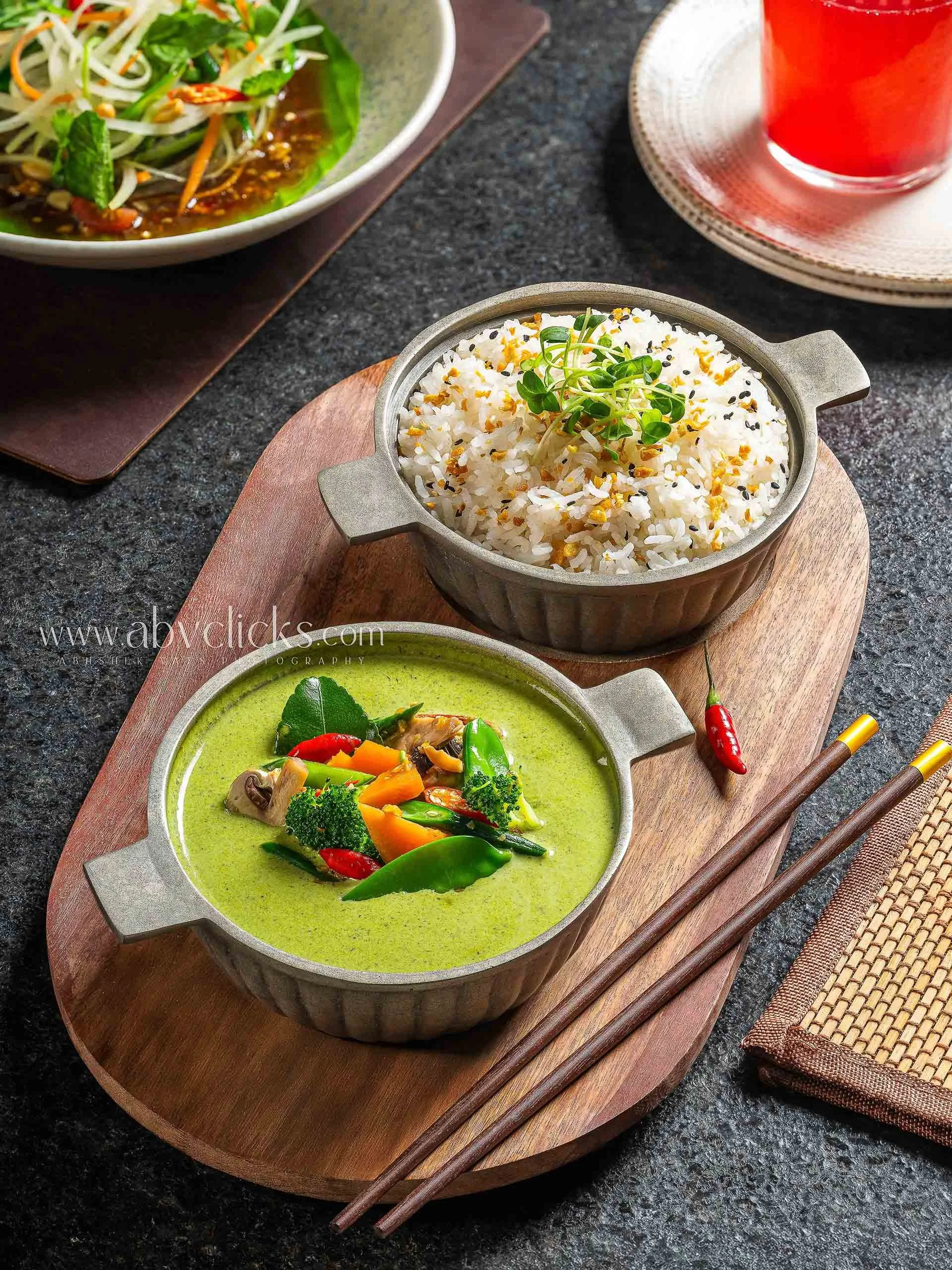 A meal with various Asian dishes, including a bowl of white rice topped with sprouts and seasoning, a green curry with vegetables, a salad with mixed vegetables and greens, chopsticks, and a glass of red beverage on a dark table.