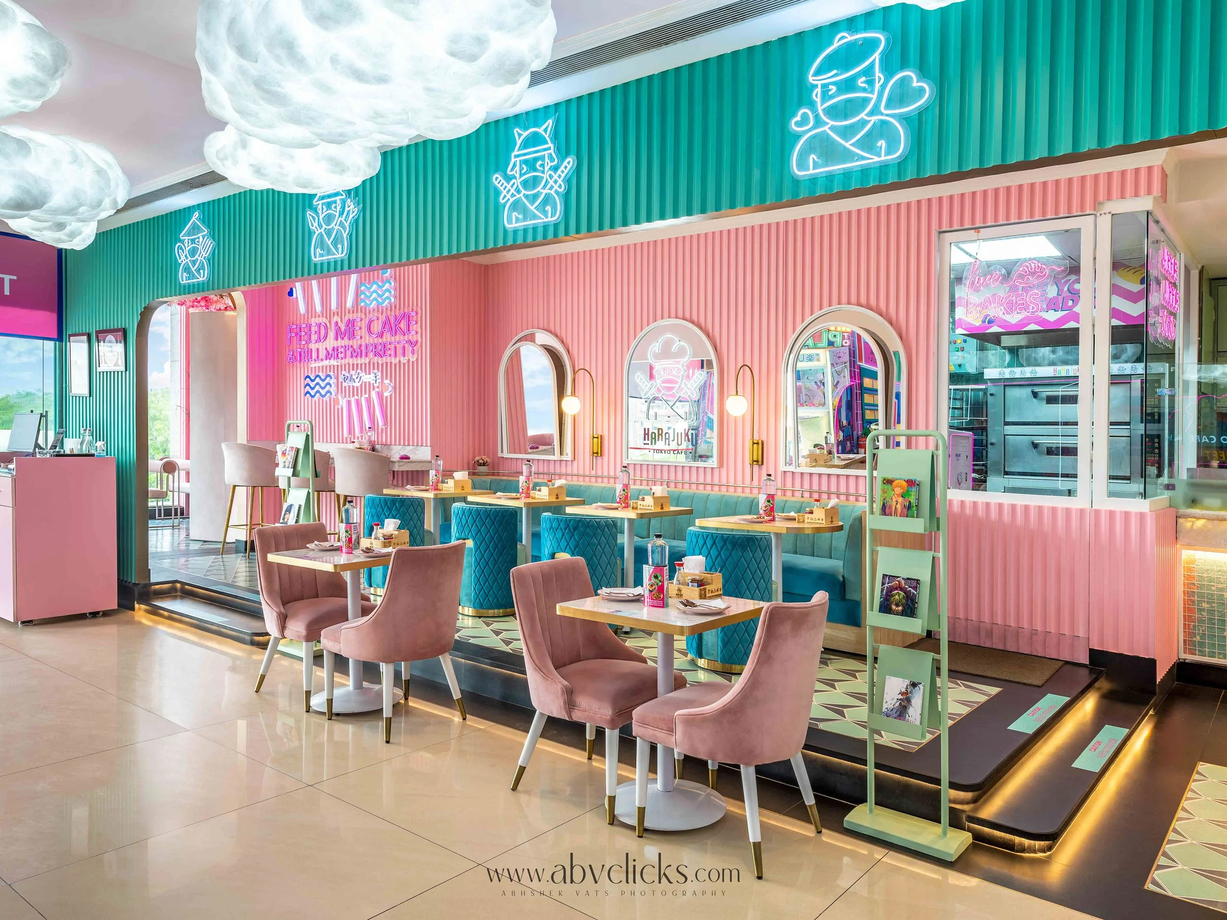 Interior of a colorful cafe with pink and teal walls decorated with neon signs and illustrations, featuring tables, chairs, and mirrors.